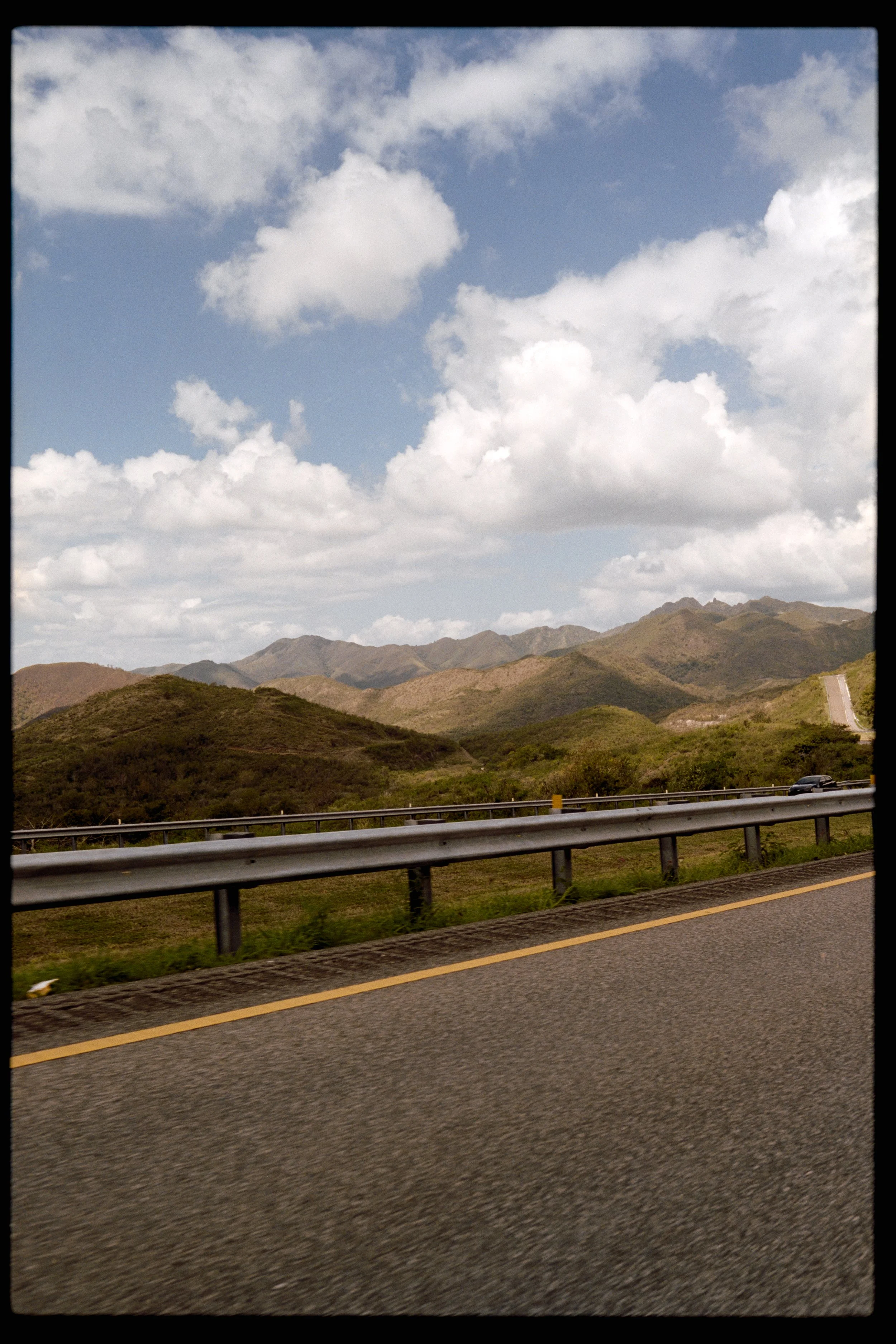 A highway with a car driving past green hills and mountains under a partly cloudy sky.