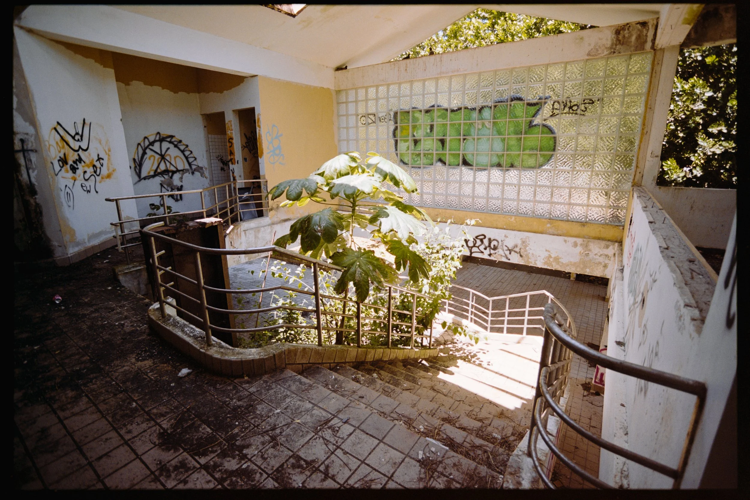 An abandoned building interior with graffiti on the walls, a broken staircase, and a large glass block window with a green landscape outside.