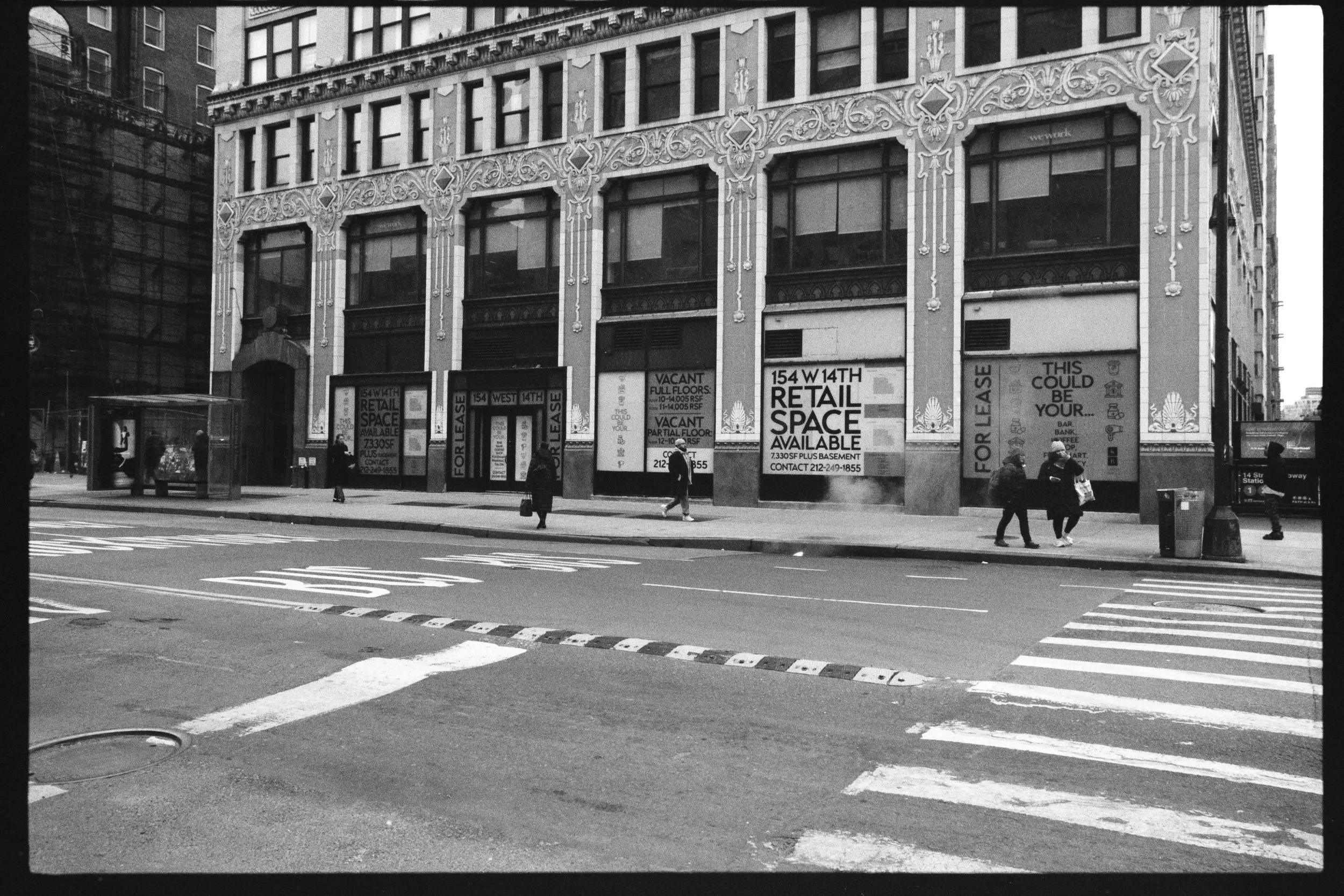 Black and white photo of a city street corner with a multi-story building featuring ornate façade details. Pedestrians are walking near the building, and storefront signs offer retail space for lease.