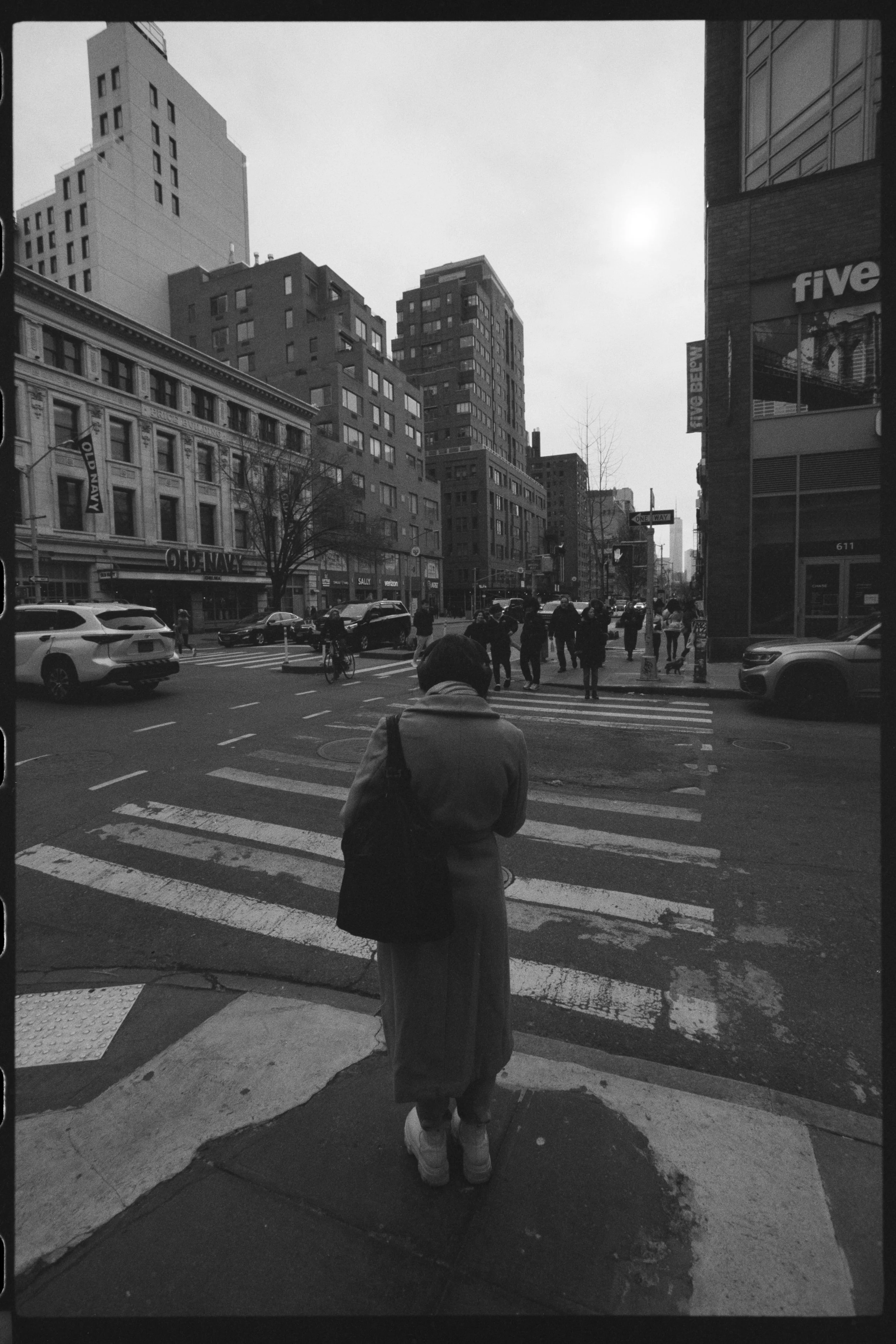 A person in a coat and sneakers is standing on a city sidewalk at a crosswalk, waiting to cross the street, with tall buildings and cars in the background.