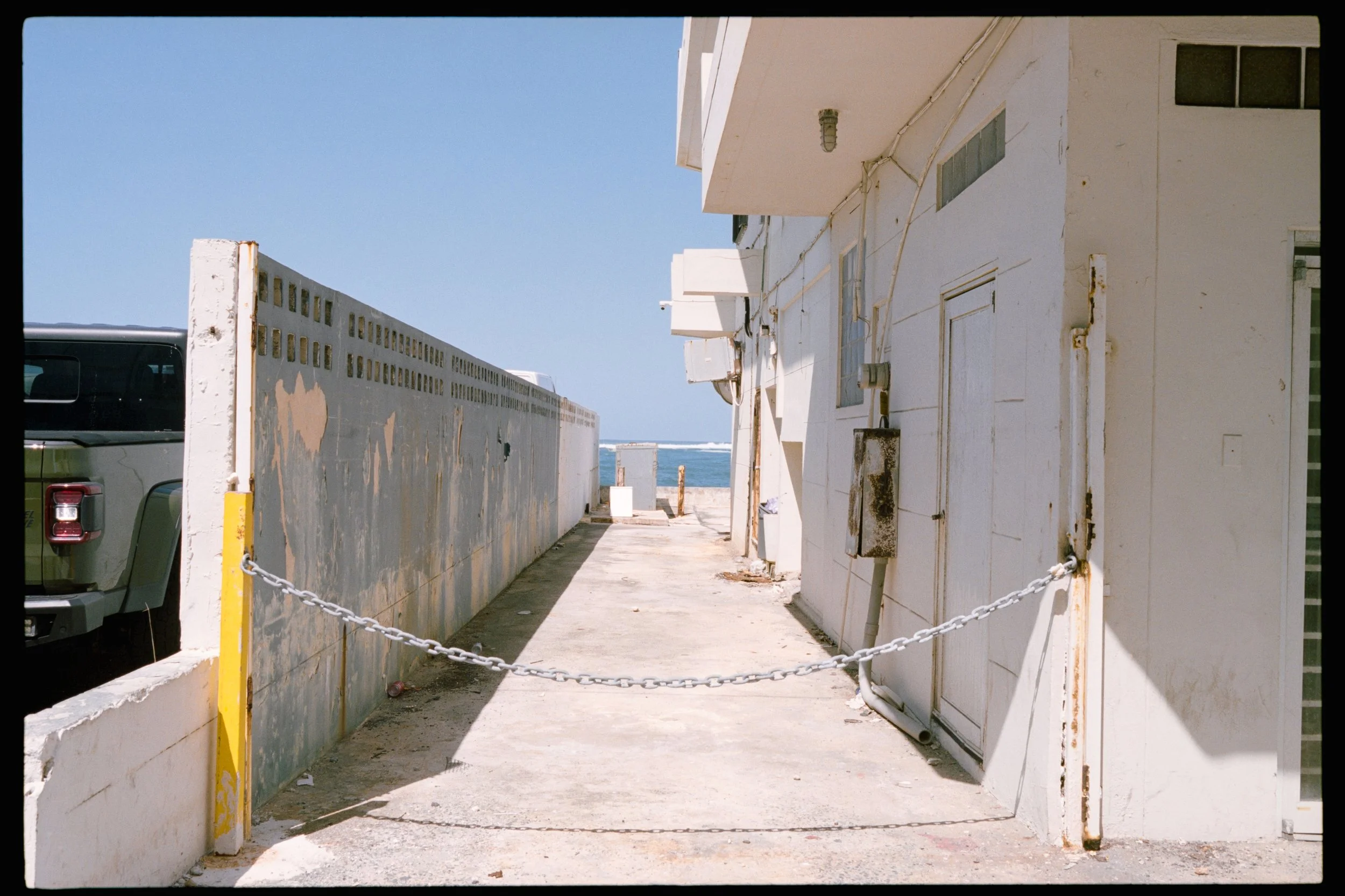 View of a narrow alley between white buildings facing the ocean, with a chain barrier and a parked black vehicle partially visible on the left.