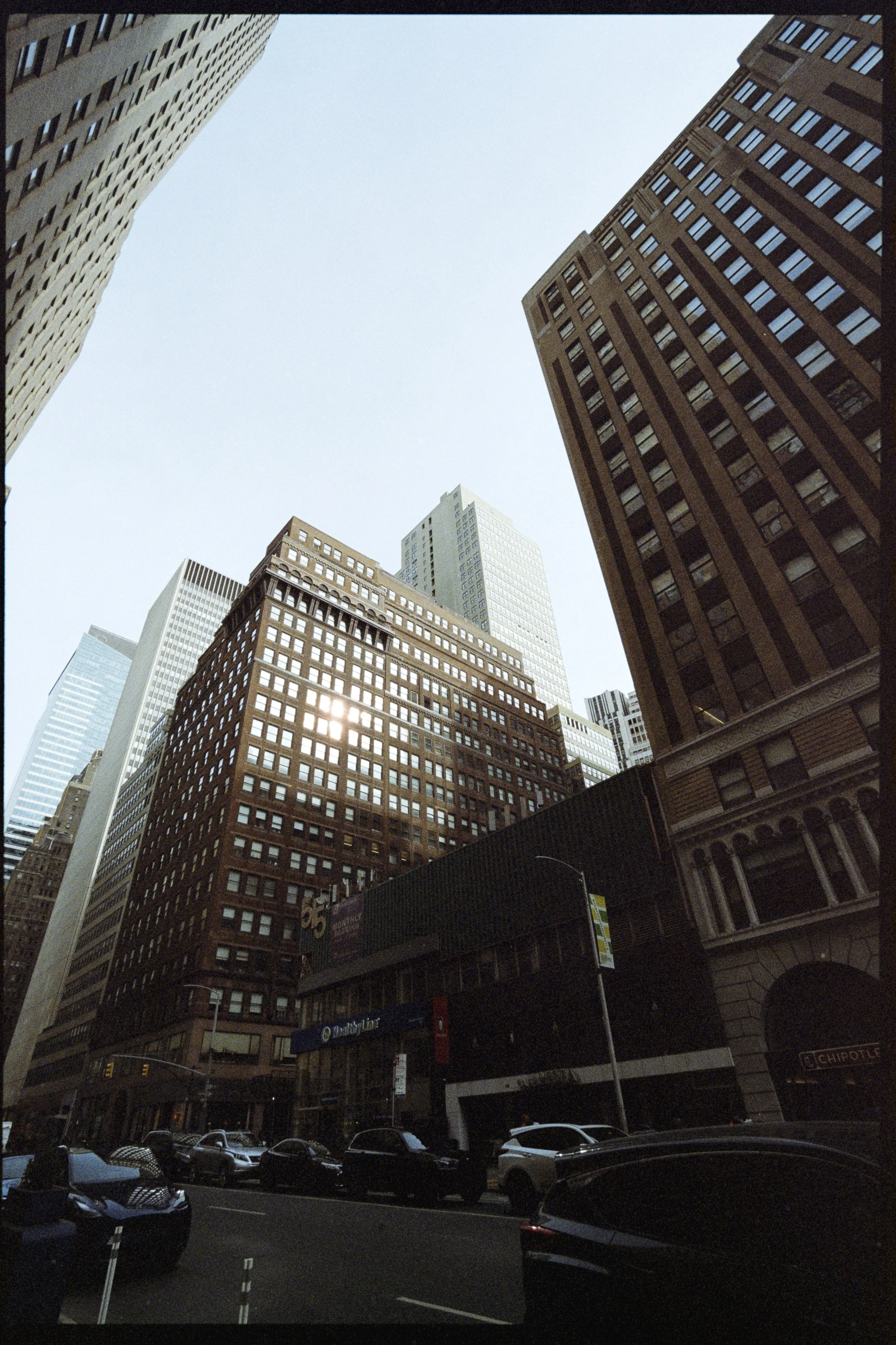 Looking up at tall skyscrapers in an urban city environment with a clear sky overhead. Multiple buildings with glass and brick facades dominate the scene, with some parked cars at street level.