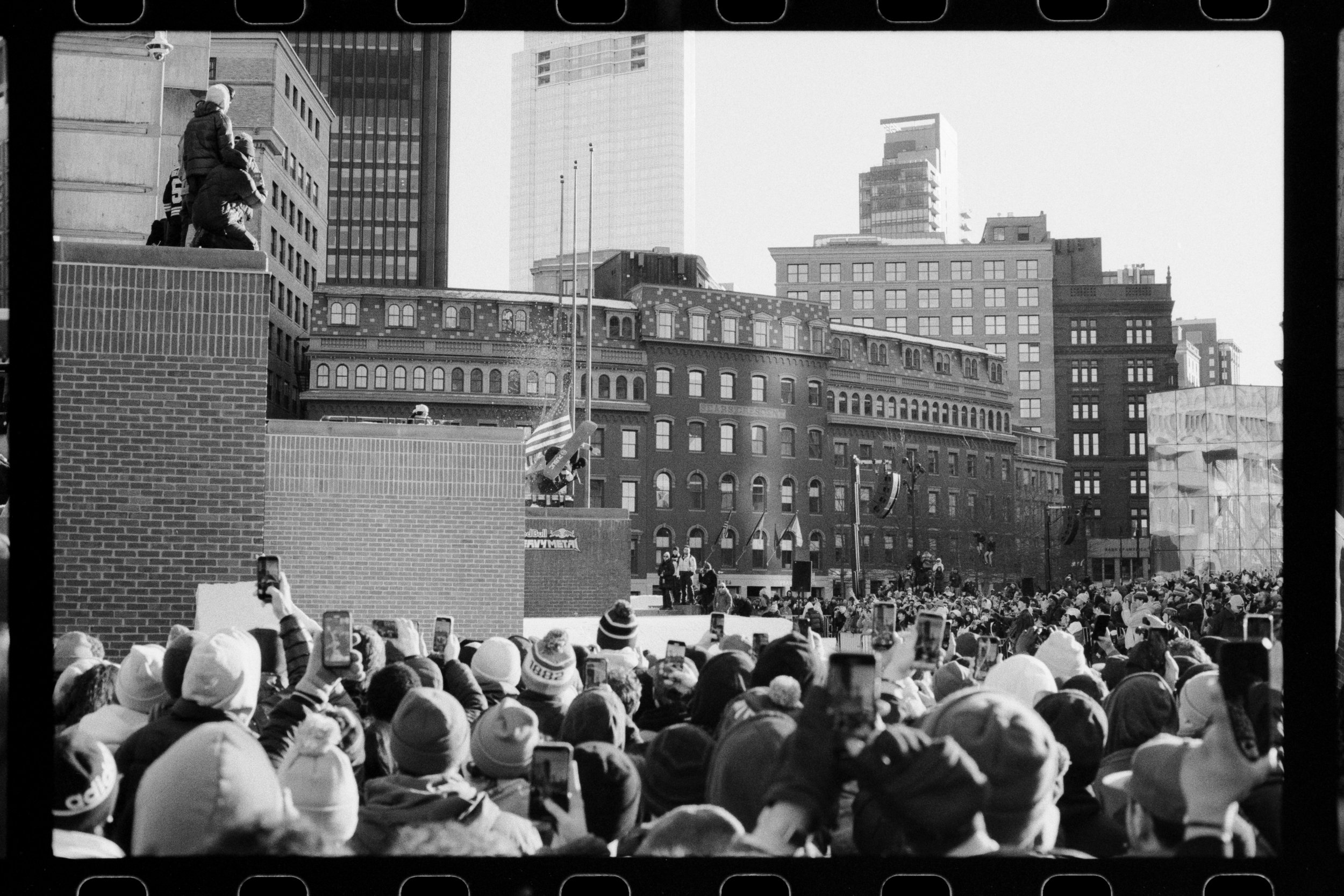 A crowd gathers at a large outdoor event in an urban setting, with many people taking photos or videos with their phones. On an elevated platform, a person is climbing with others, possibly for a stunt or performance, against a backdrop of tall skysc