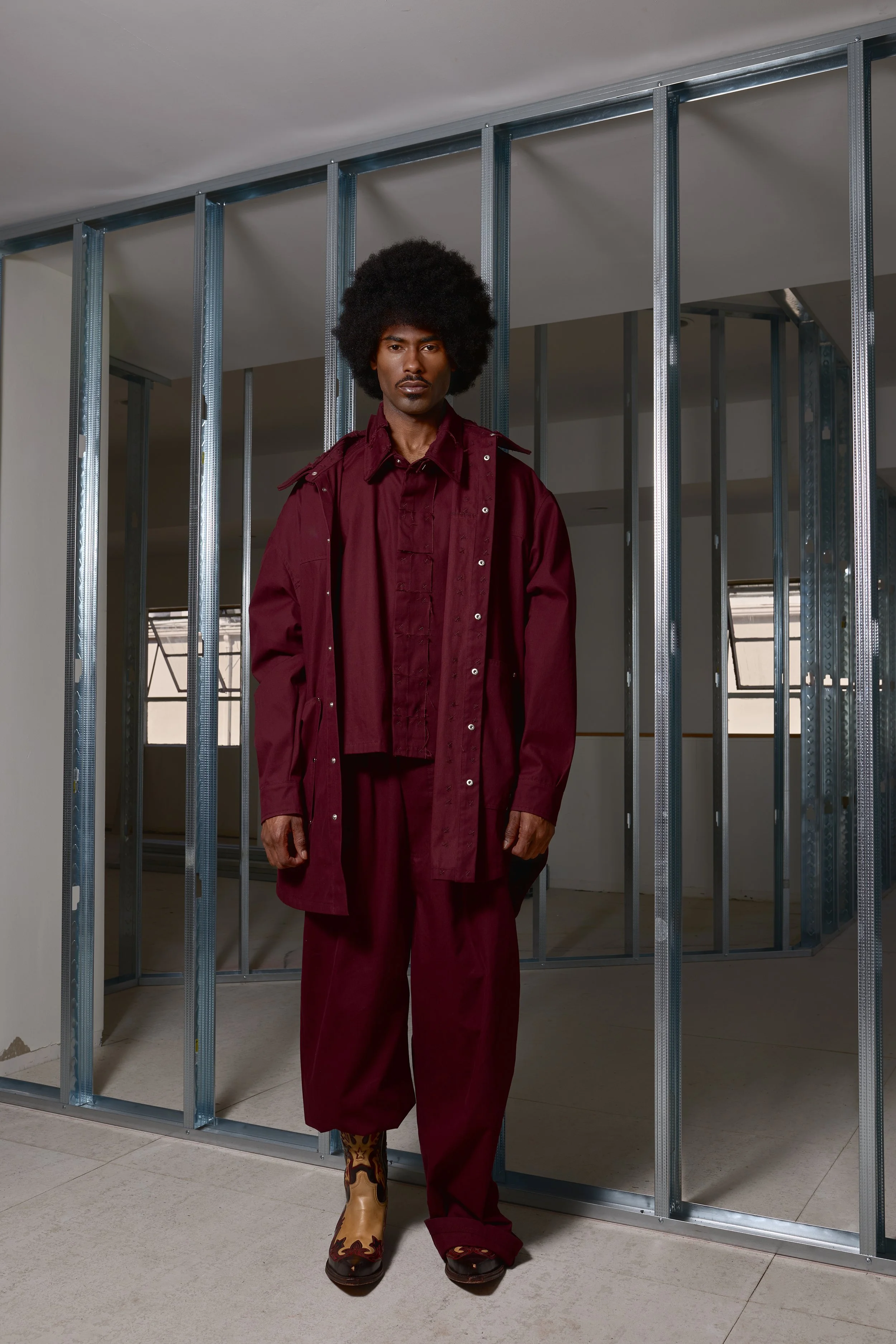 A man with an afro hairstyle wearing a maroon outfit and cowboy boots standing inside a room with metal framing for walls under construction.