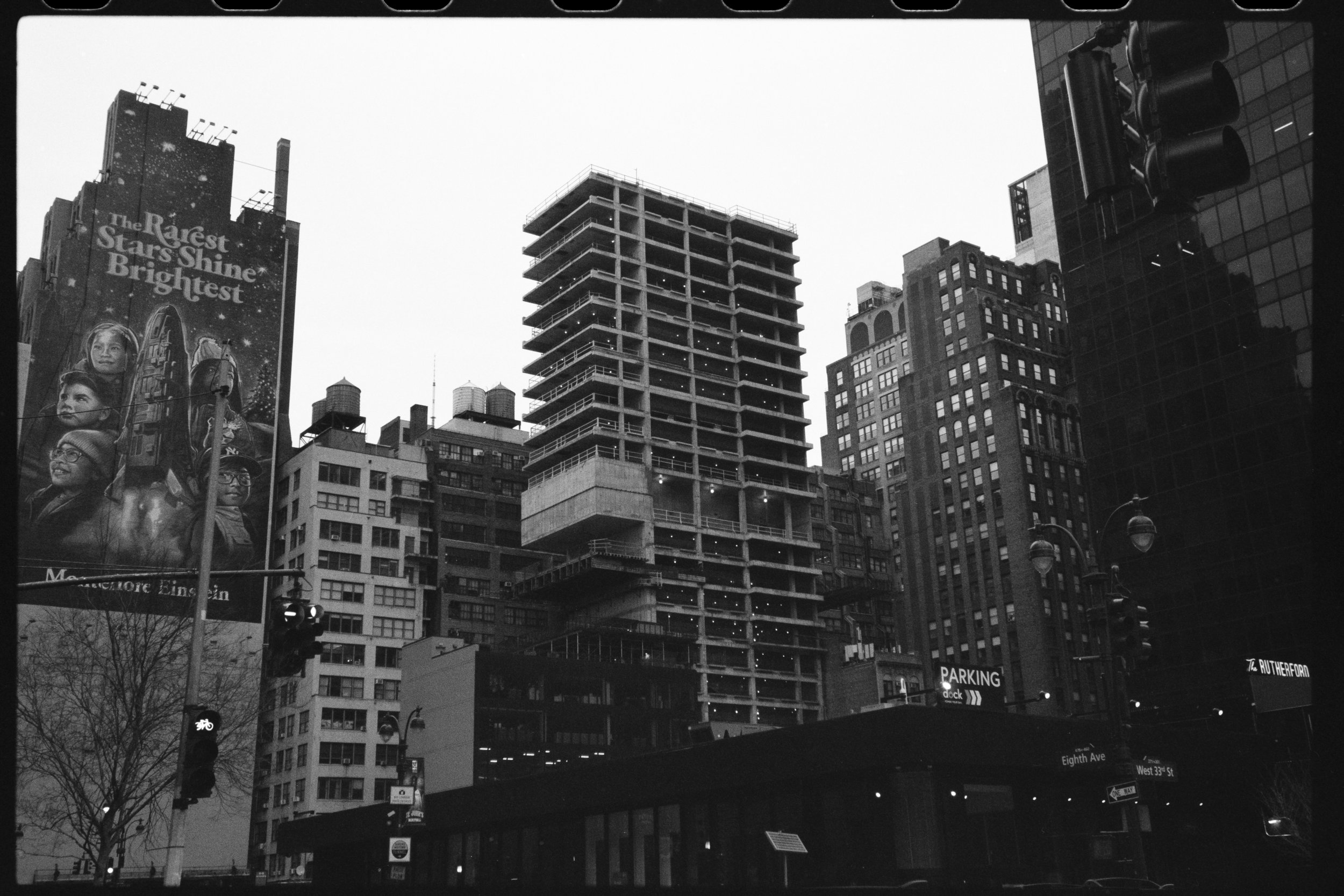 Black and white photo of a cityscape with tall buildings, including an unfinished high-rise, a billboard with the message 'The Rarest Stars Shine Brightest,' and traffic lights at an intersection.