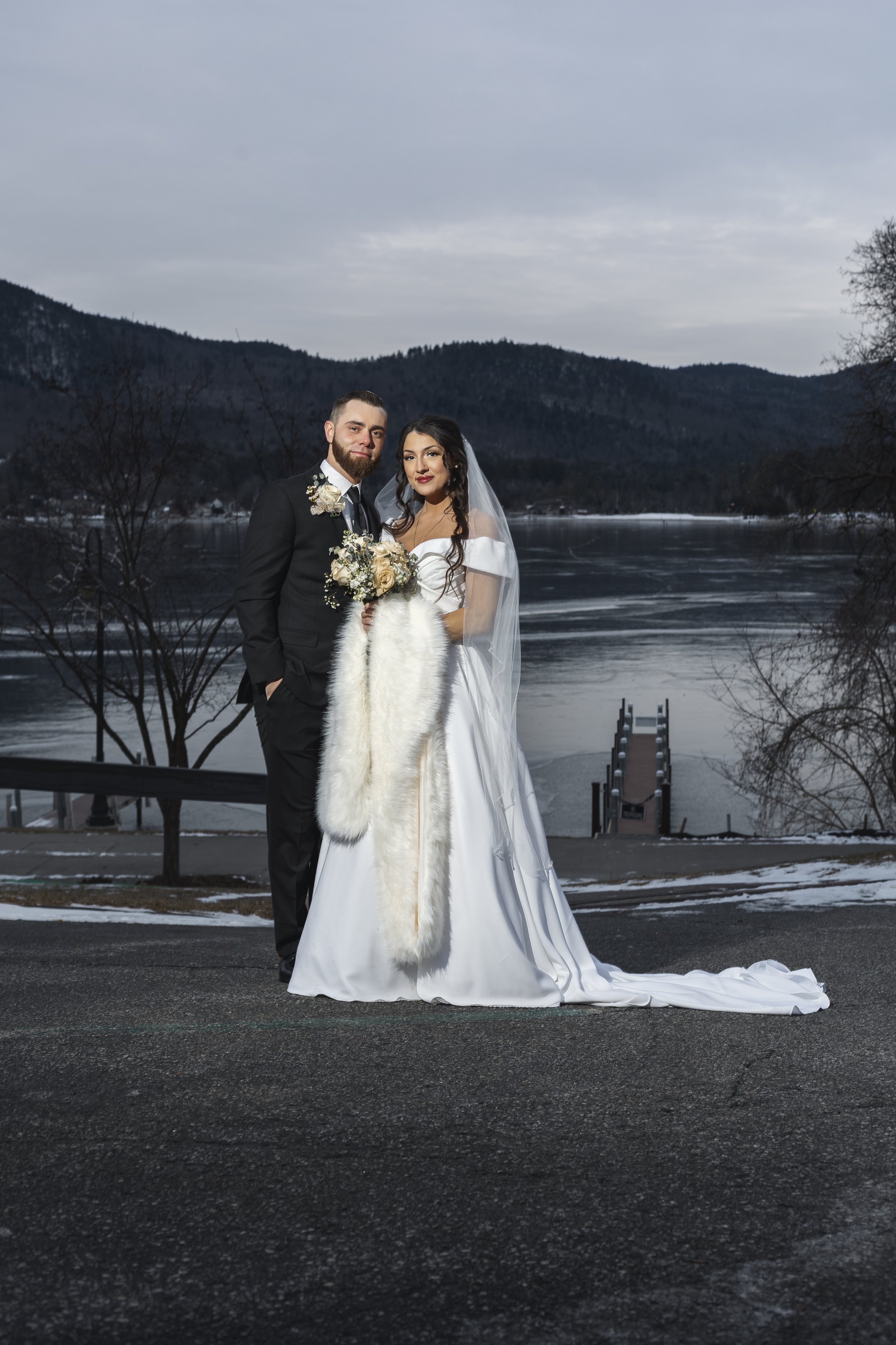 A newlywed couple stand outdoors on a paved road near a lake in winter, with mountains in the background. The bride is wearing a white wedding gown and a veil, holding a bouquet, with a white fur stole. The groom is dressed in a black suit with a bou