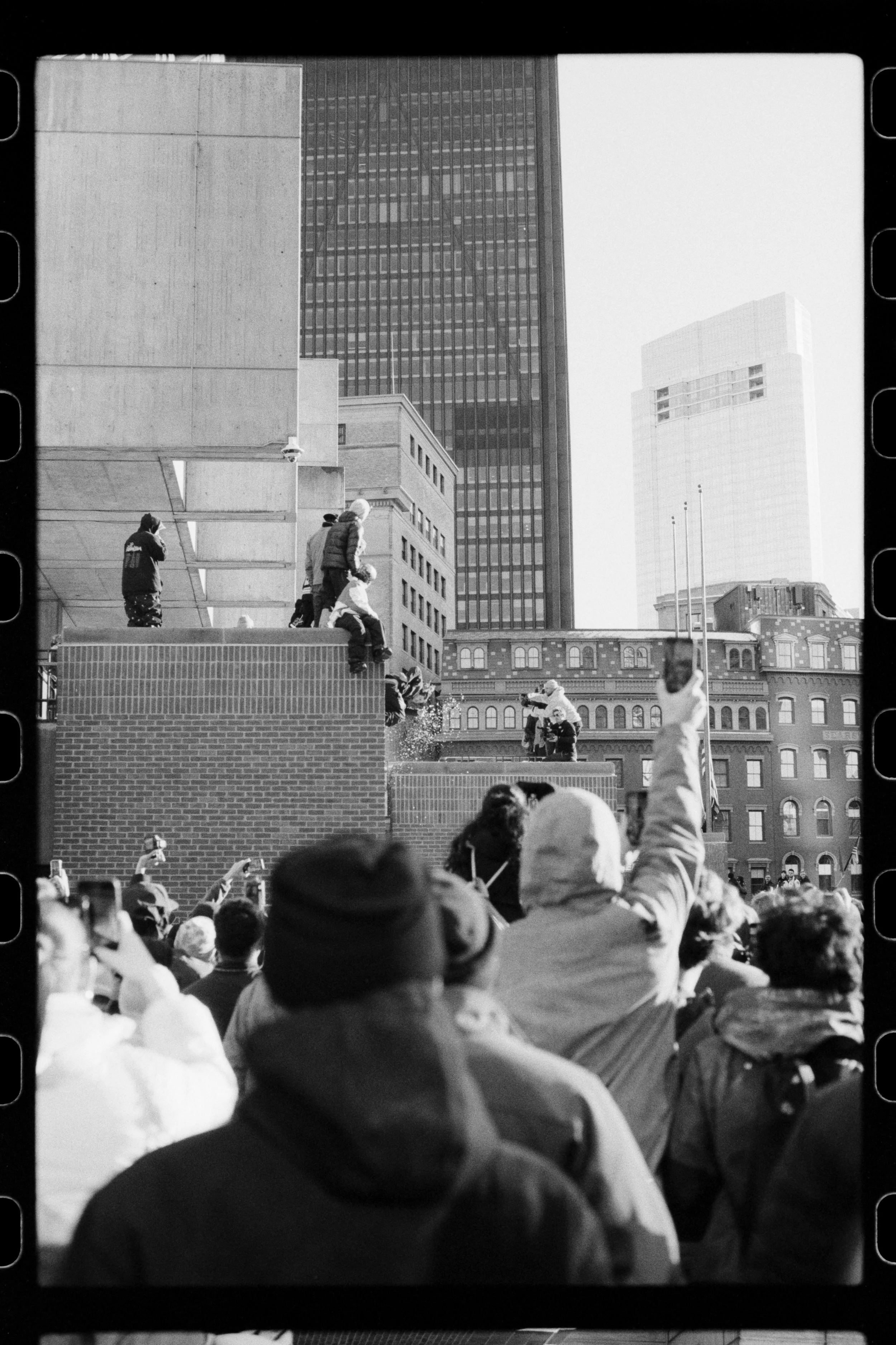 A crowd gathered in an urban area with tall modern and older buildings in the background. Some people are taking photos or recording videos, and others are sitting on a ledge or walking around.