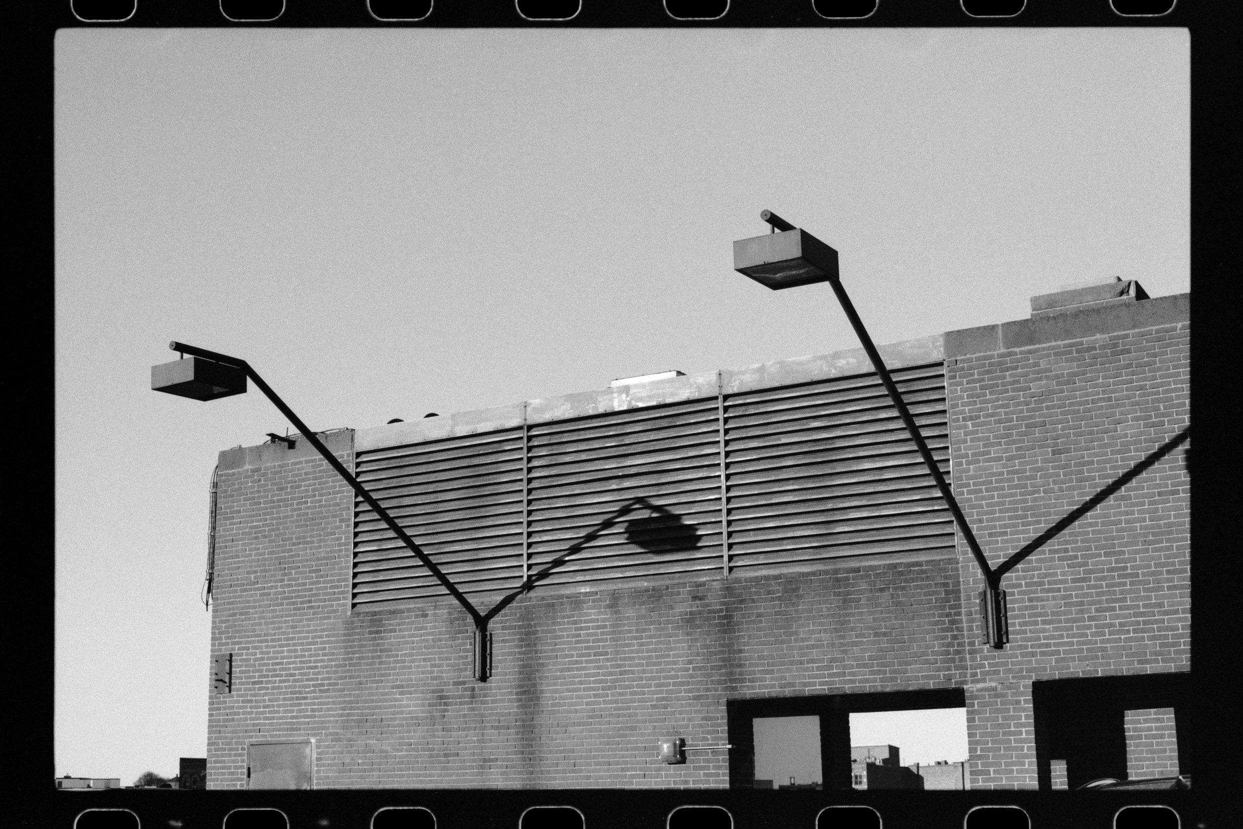 Black and white photo of a brick building with a wooden fence and two streetlights, shadows cast on the wall, clear sky in the background.