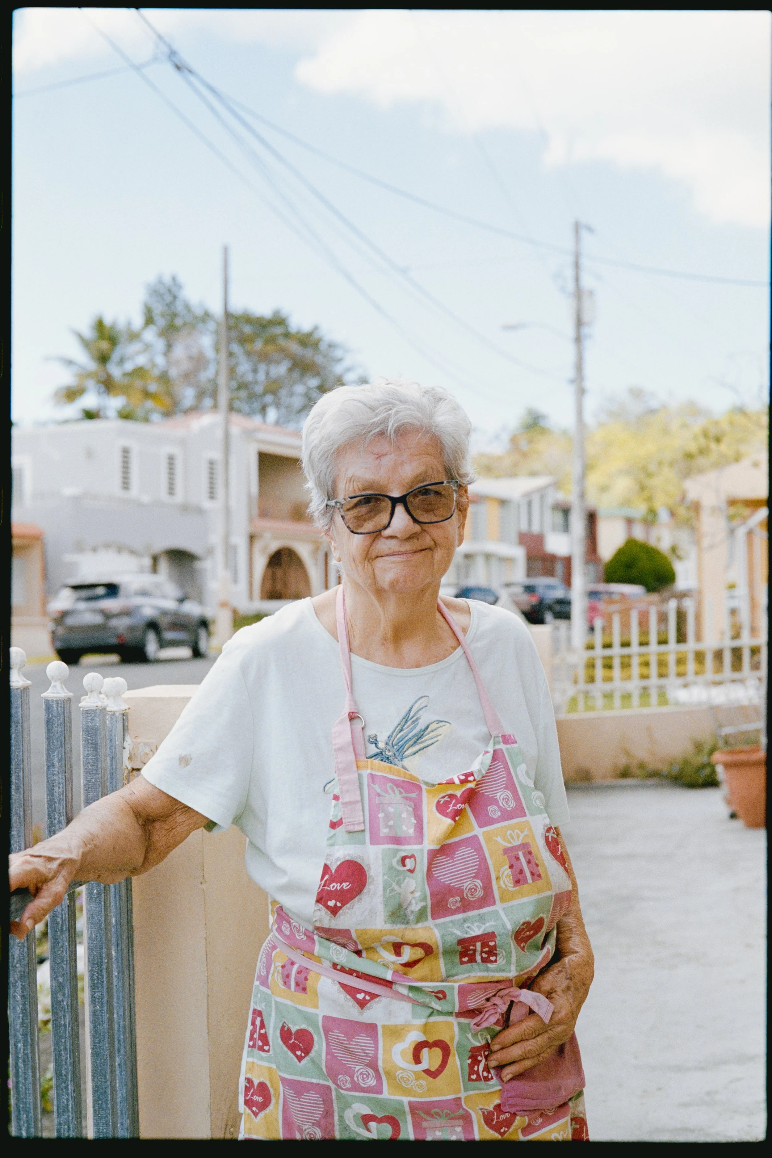 An elderly woman with white hair, wearing glasses and a colorful apron with various love-themed symbols, stands outdoors on a sunny day in a residential neighborhood. She is smiling and holding onto a fence with one arm.