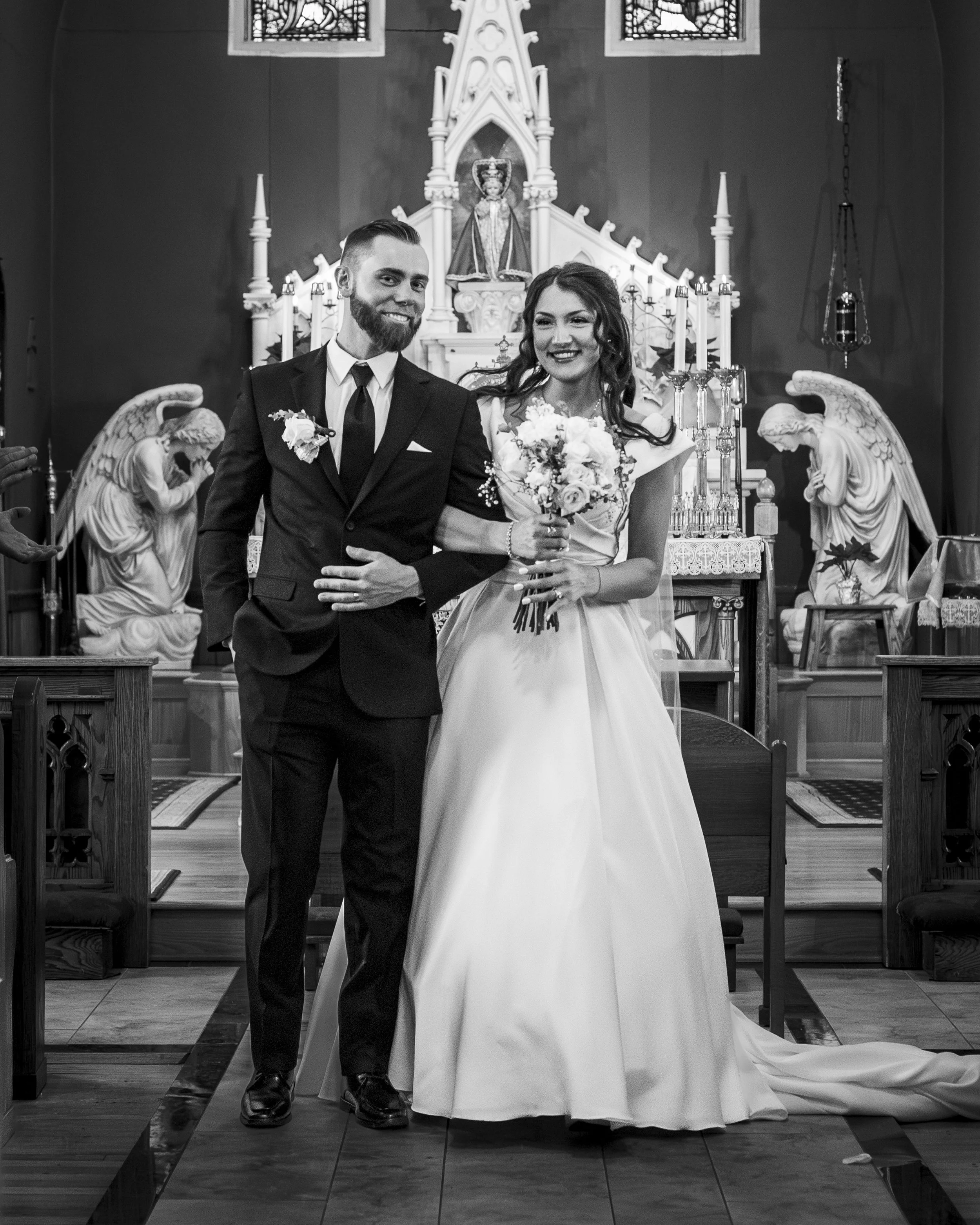 A black and white photo of a bride and groom standing in a church, smiling. The groom is wearing a dark suit with a tie, and the bride is wearing a wedding dress and holding a bouquet of flowers. The church interior features statues, stained glass wi