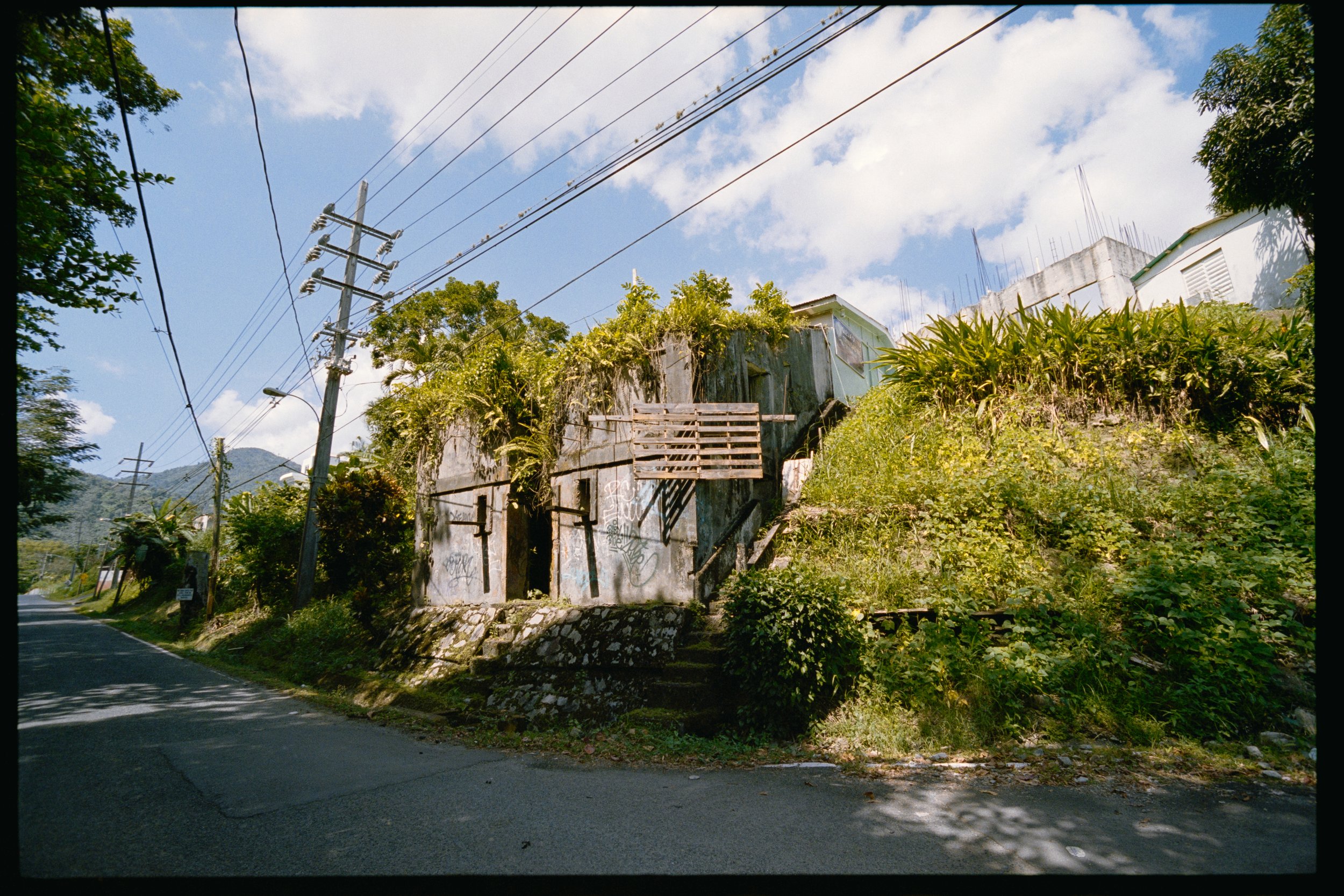 A hillside scene with an old abandoned building, surrounded by lush green vegetation and power lines overhead.