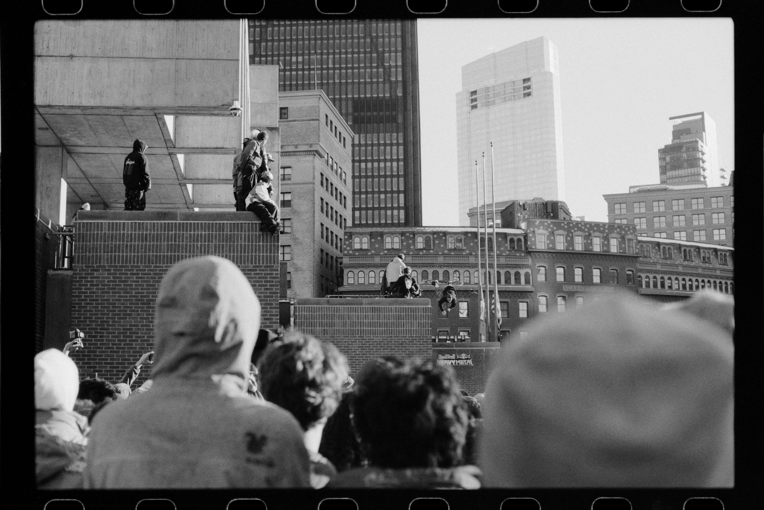 Crowd of people watching a performance or event in an urban outdoor setting with tall buildings in the background.