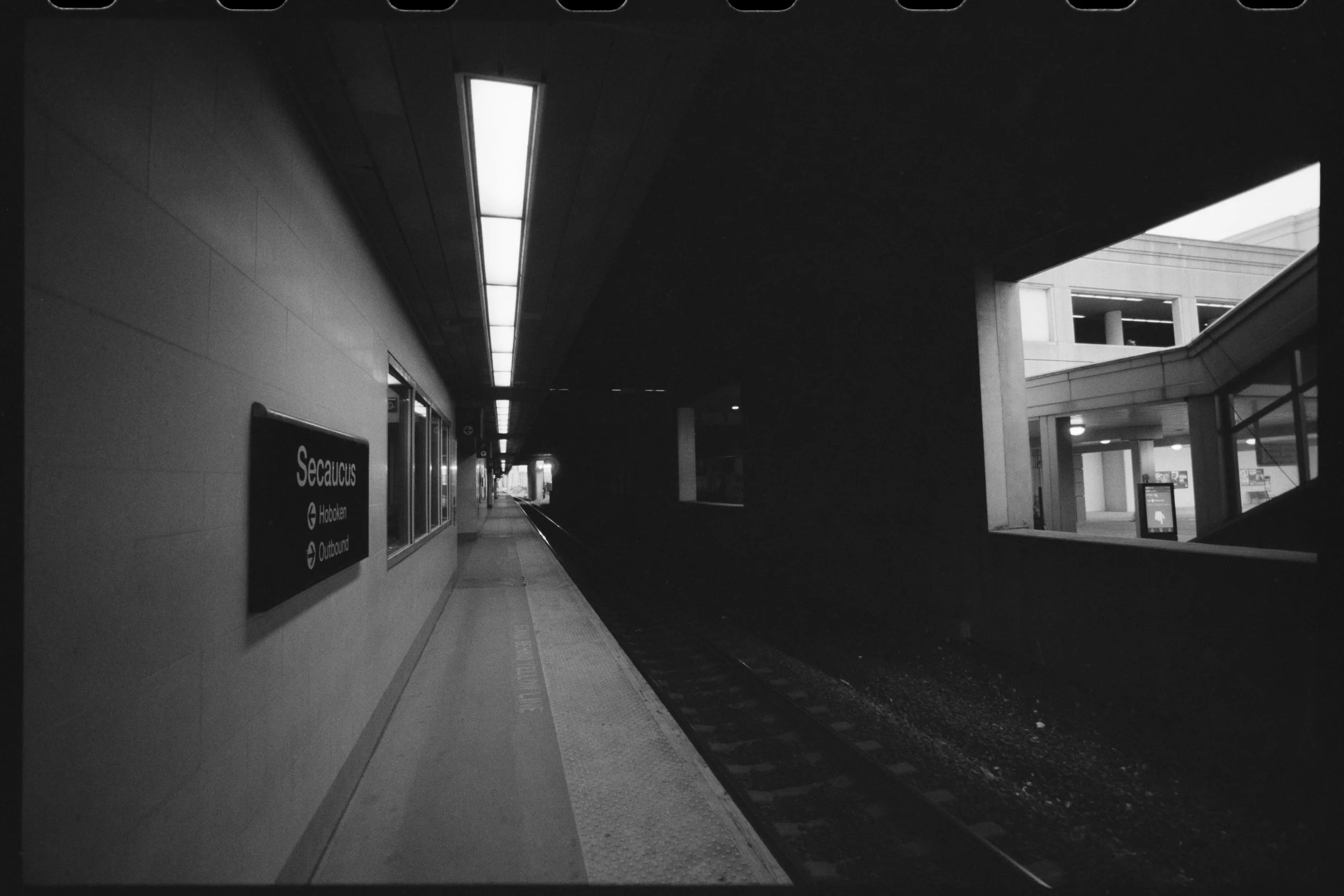 An underground train station platform with a sign indicating directions to Secaucus, Hoboken, and Outbound. The platform is empty with a modern design, featuring a long row of ceiling lights, and modern architecture visible in the background.