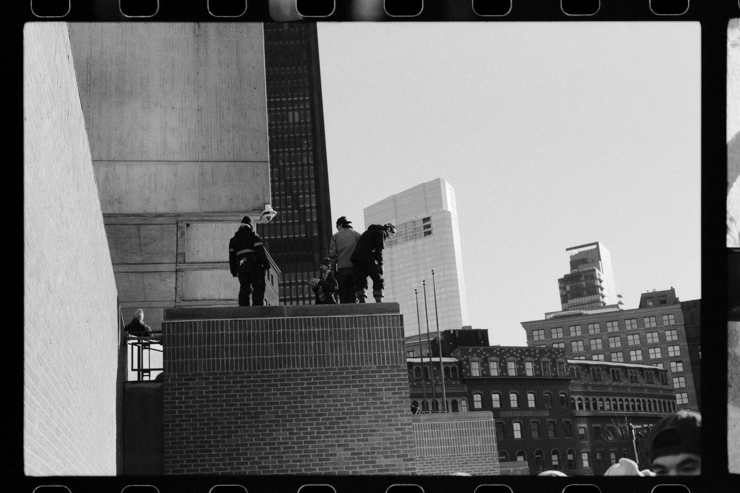 Black and white photo of people walking and sitting on a brick ledge on a city street with tall buildings in the background.