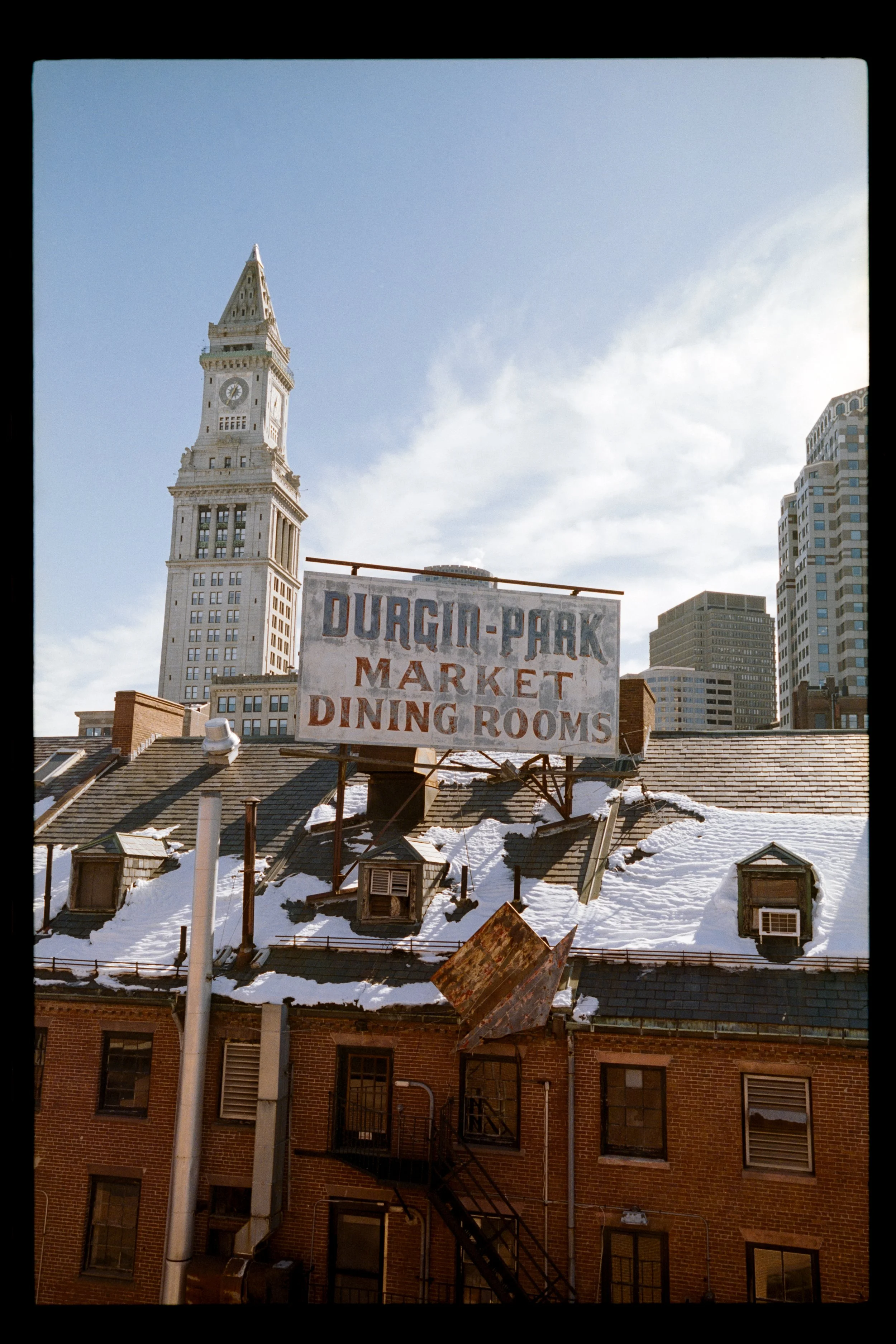 View of the San Francisco cityscape featuring the iconic Clock Tower building, with a sign for the Durgin-Park Market and Dining Rooms in the foreground of snowy rooftops and brick buildings.