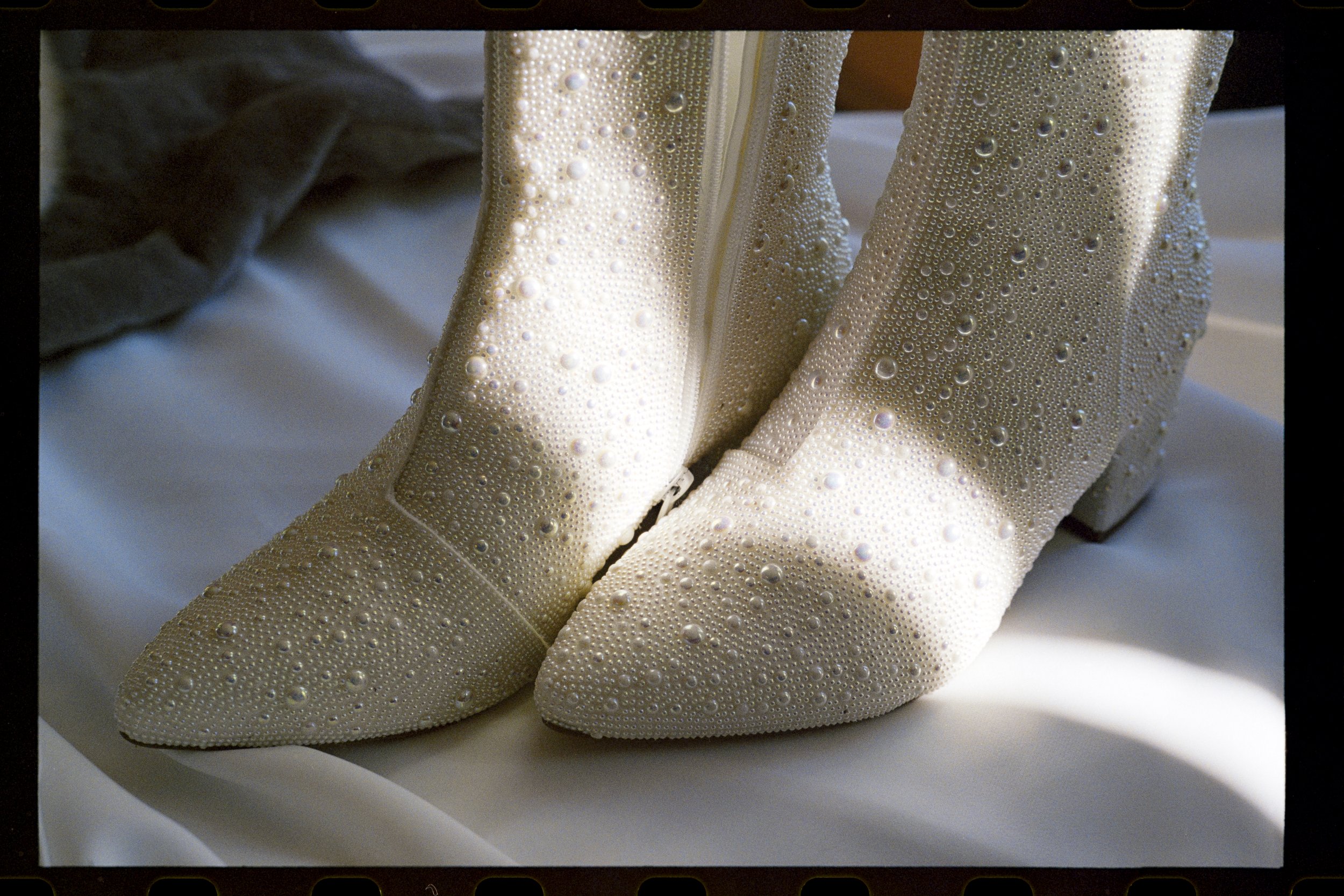 Close-up of a pair of beige high-heeled shoes decorated with numerous small, sparkling rhinestones, displayed on a white surface.