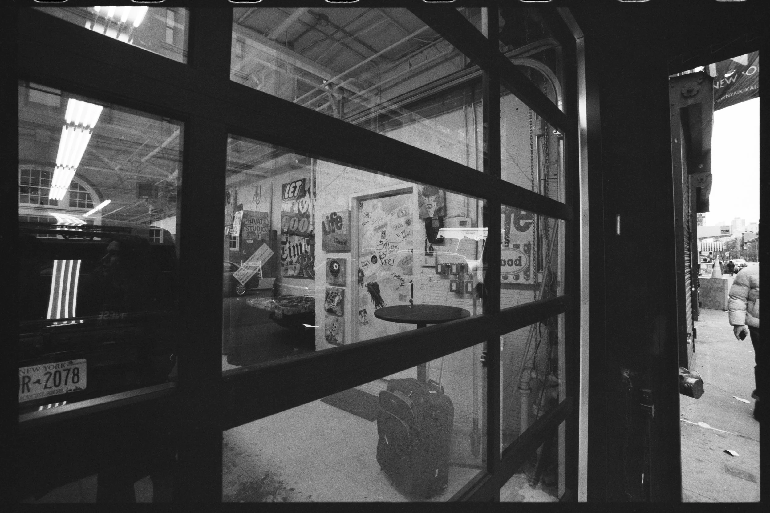 View through a glass window of a street scene with a person walking on the sidewalk, several signs, and part of a building with advertisements, all in black and white.