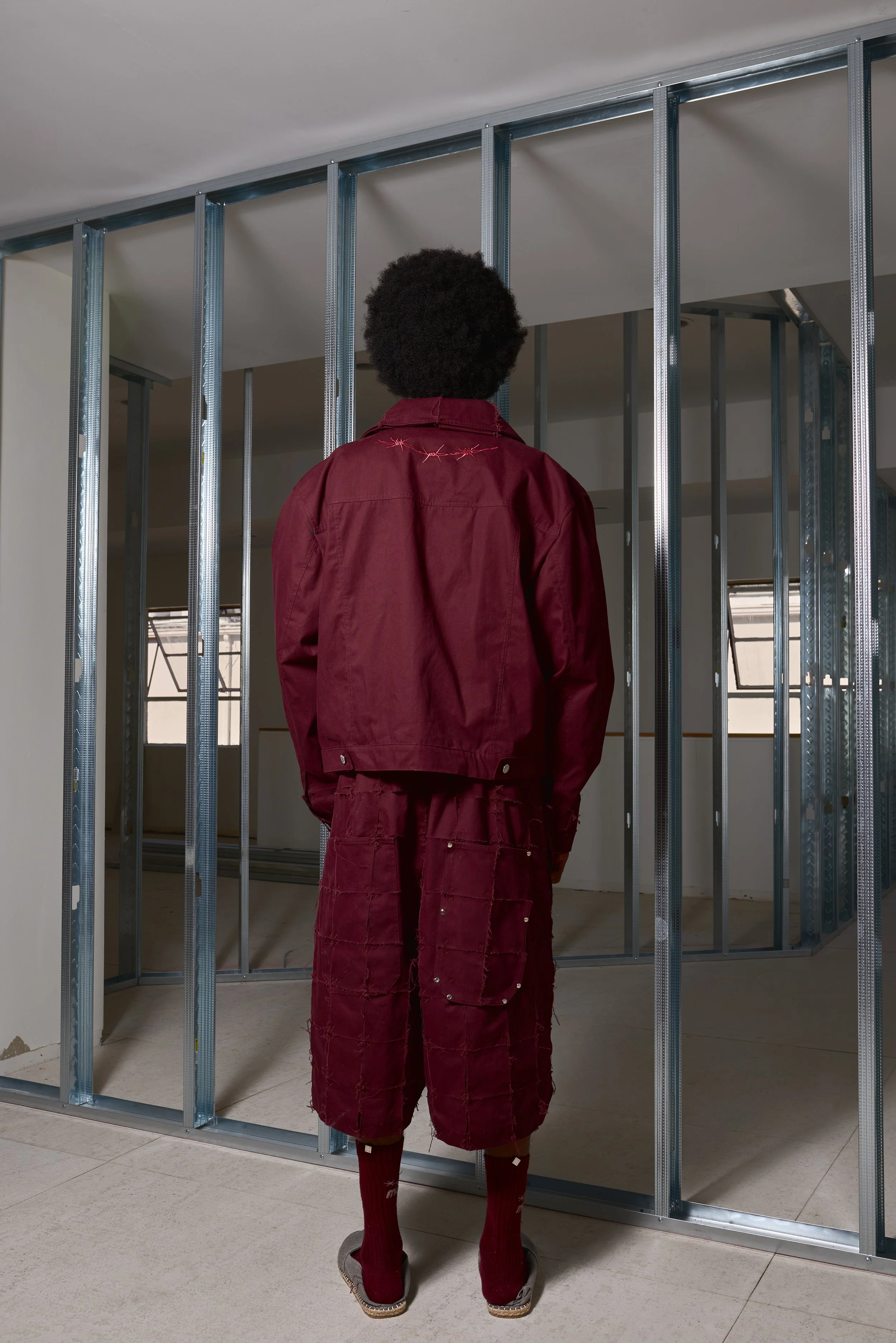 Person with curly hair wearing a maroon jacket and shorts, standing inside an unfinished room with exposed metal framing.