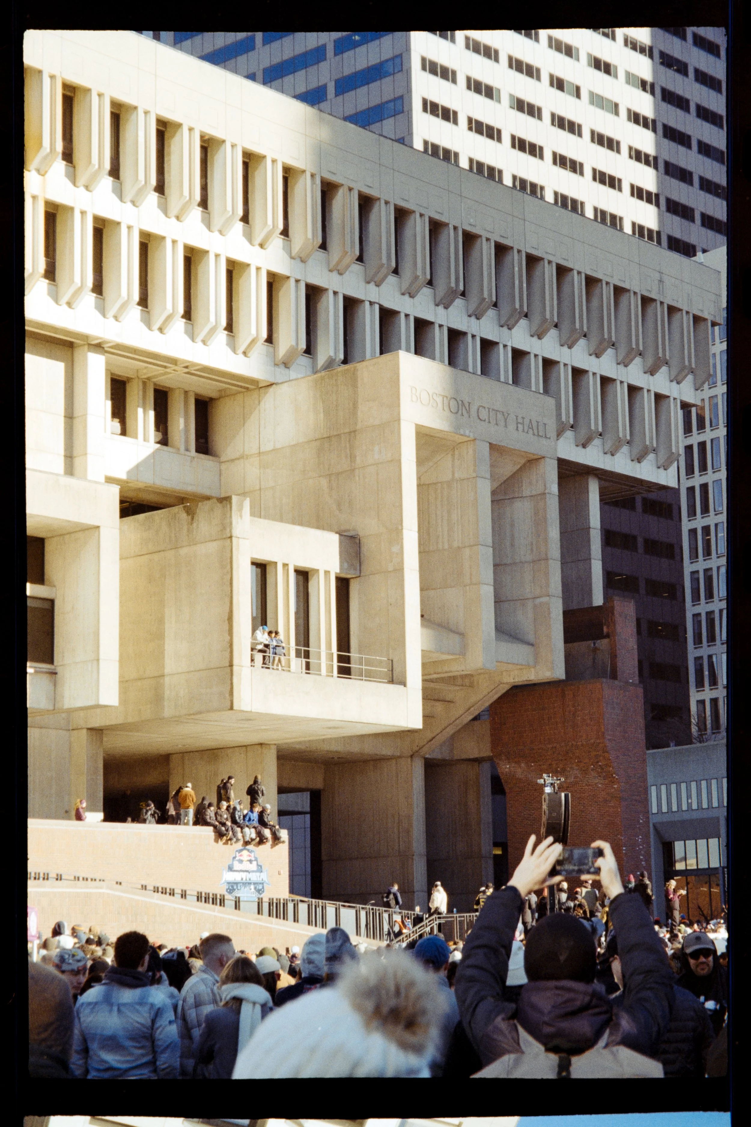 Crowd gathered outside Boston City Hall, with some people taking photos. The building has a modern architectural design with a beige facade and prominent windows. Tall skyscrapers are visible in the background.