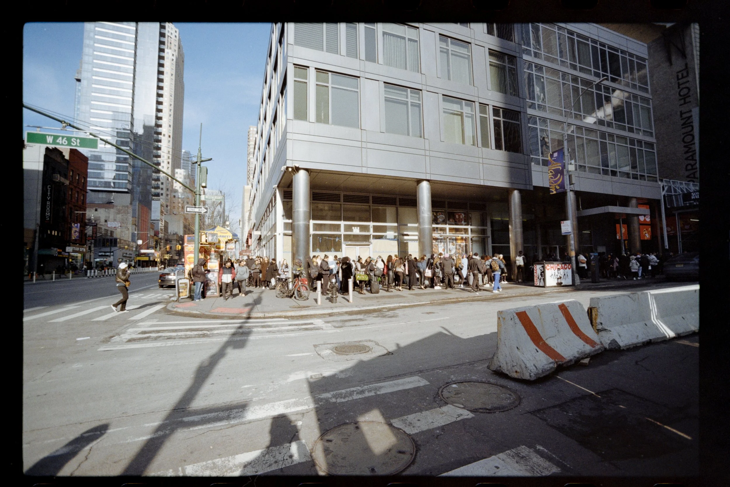 Street scene with a crowd of people waiting at a crosswalk in front of a modern glass building, with tall skyscrapers in the background, and street signs indicating W 46 St.