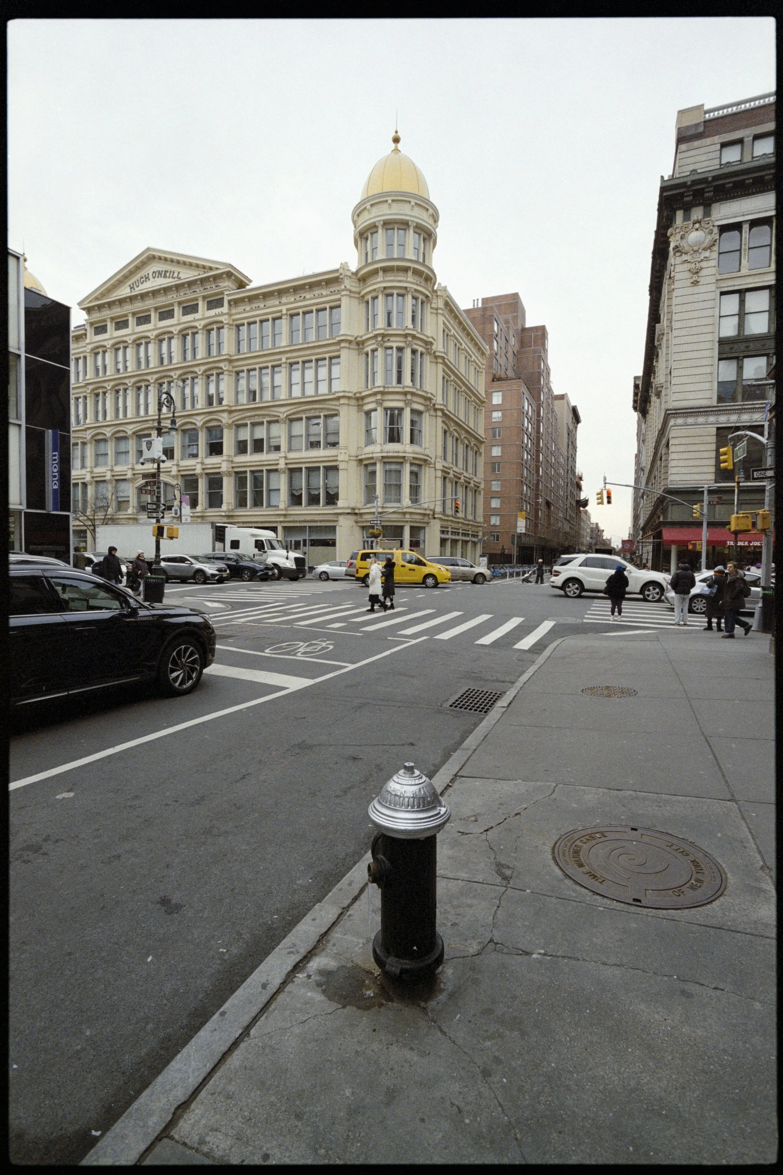 City street scene with crosswalk, parked cars, pedestrians, and large historic building with a rounded corner tower topped with a golden dome.
