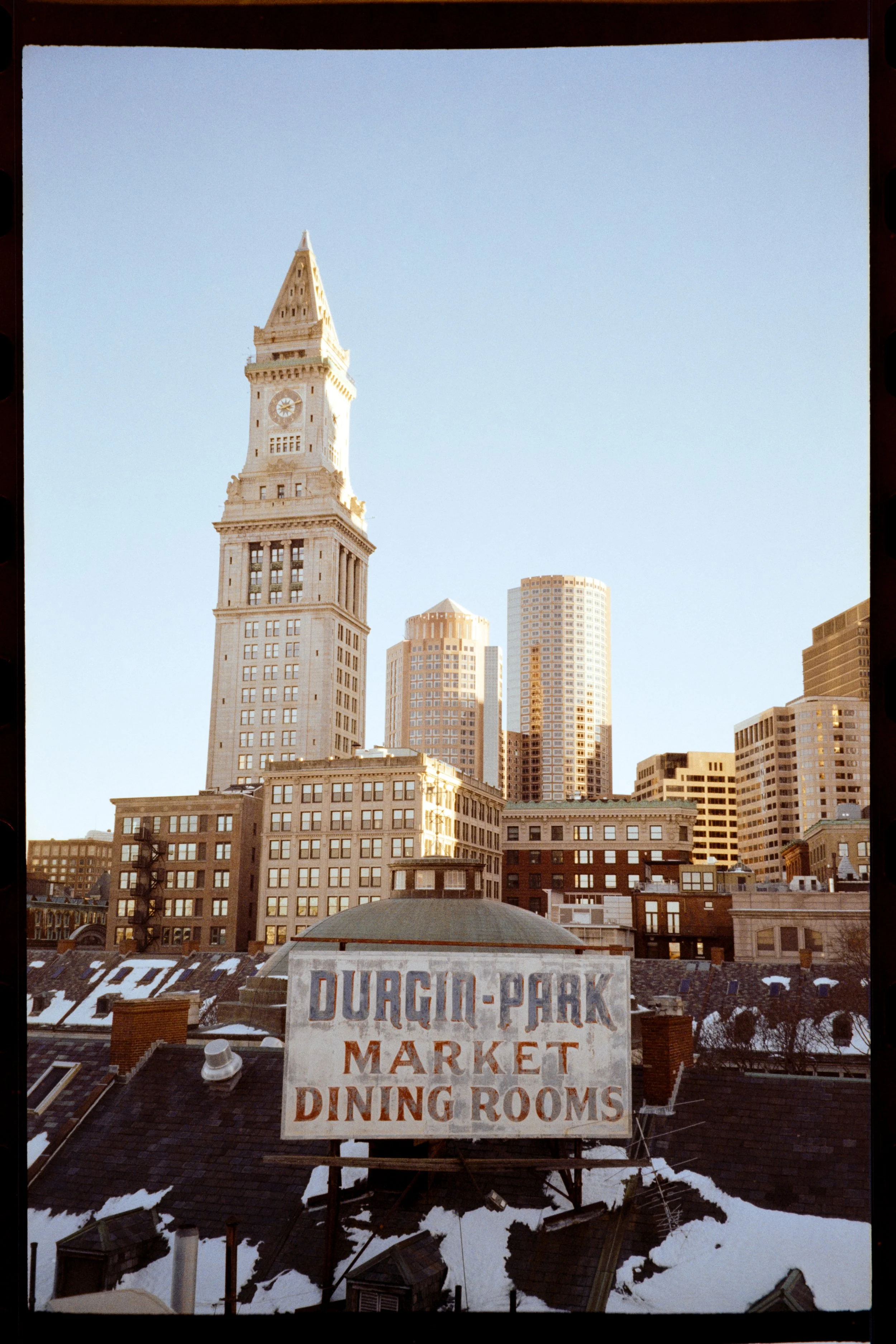 City skyline with historic clock tower and modern skyscrapers, snow-covered rooftops, and a sign that reads 'Durgin-Park Market Dining Rooms'.