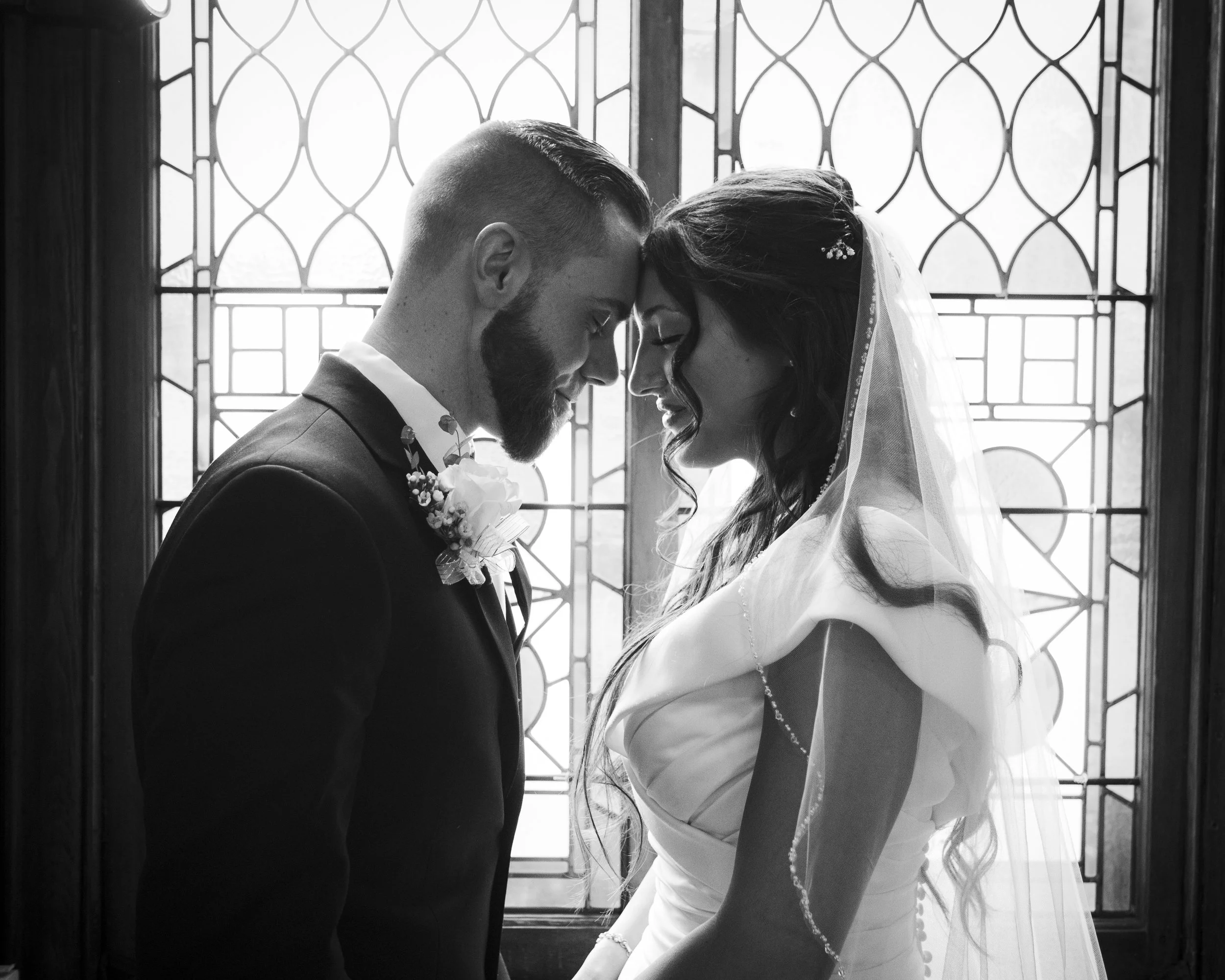 A black-and-white photo of a bride and groom with their foreheads touching, standing in front of a decorative window with stained glass, during their wedding.