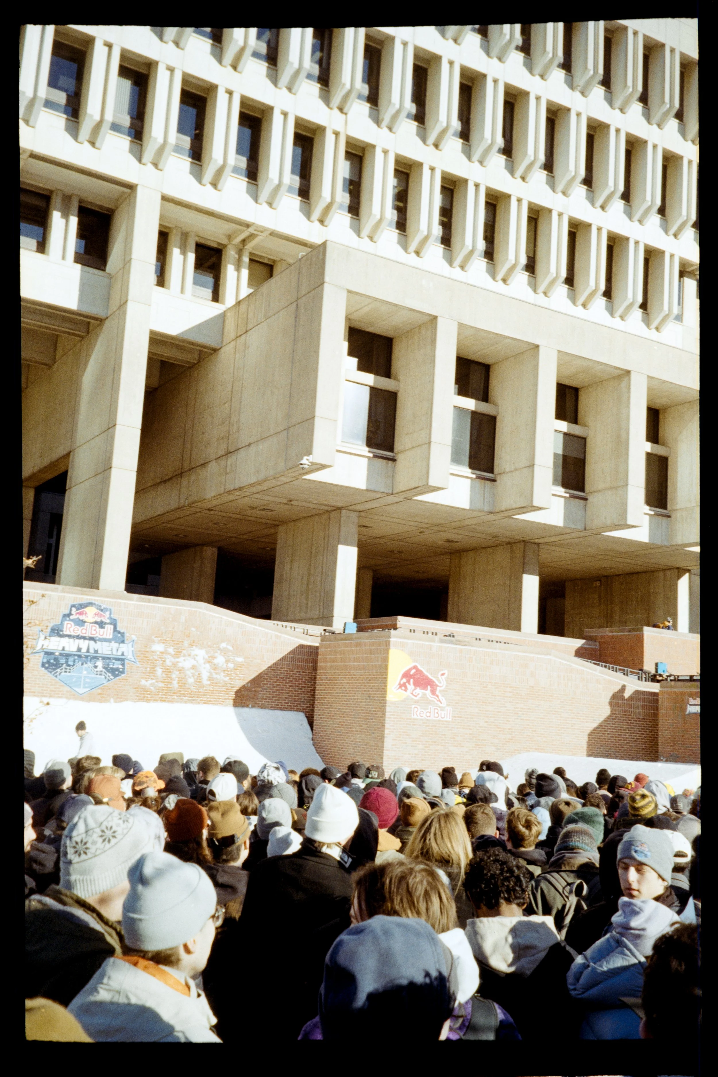 Large crowd of people wearing hats and jackets gathers outdoors in front of a building with a Red Bull Heavymetal logo on a brick wall. The building has a modern architectural style with beige concrete and multiple windows.