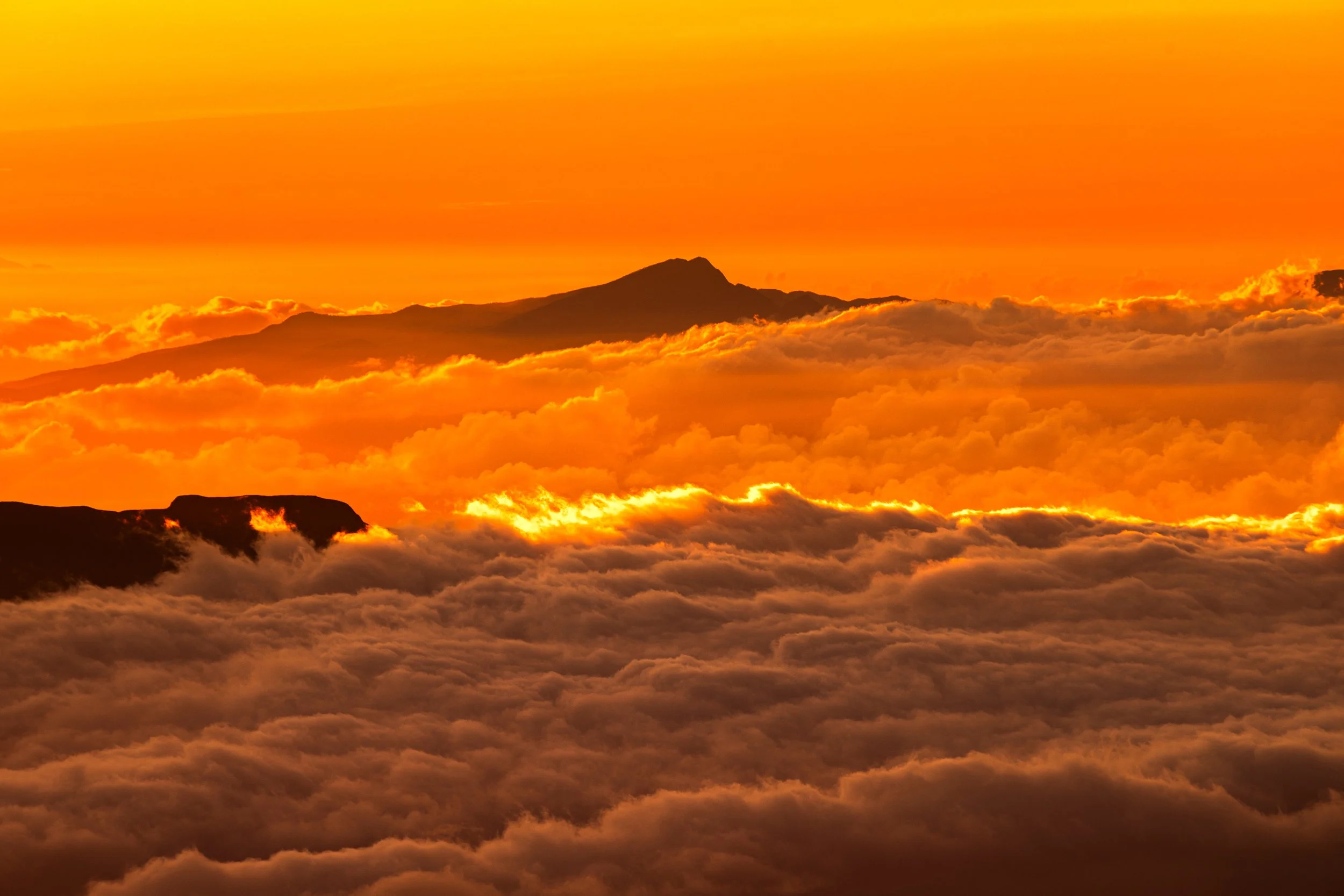 A view of a mountain surrounded by clouds at sunset with a vibrant orange and yellow sky.