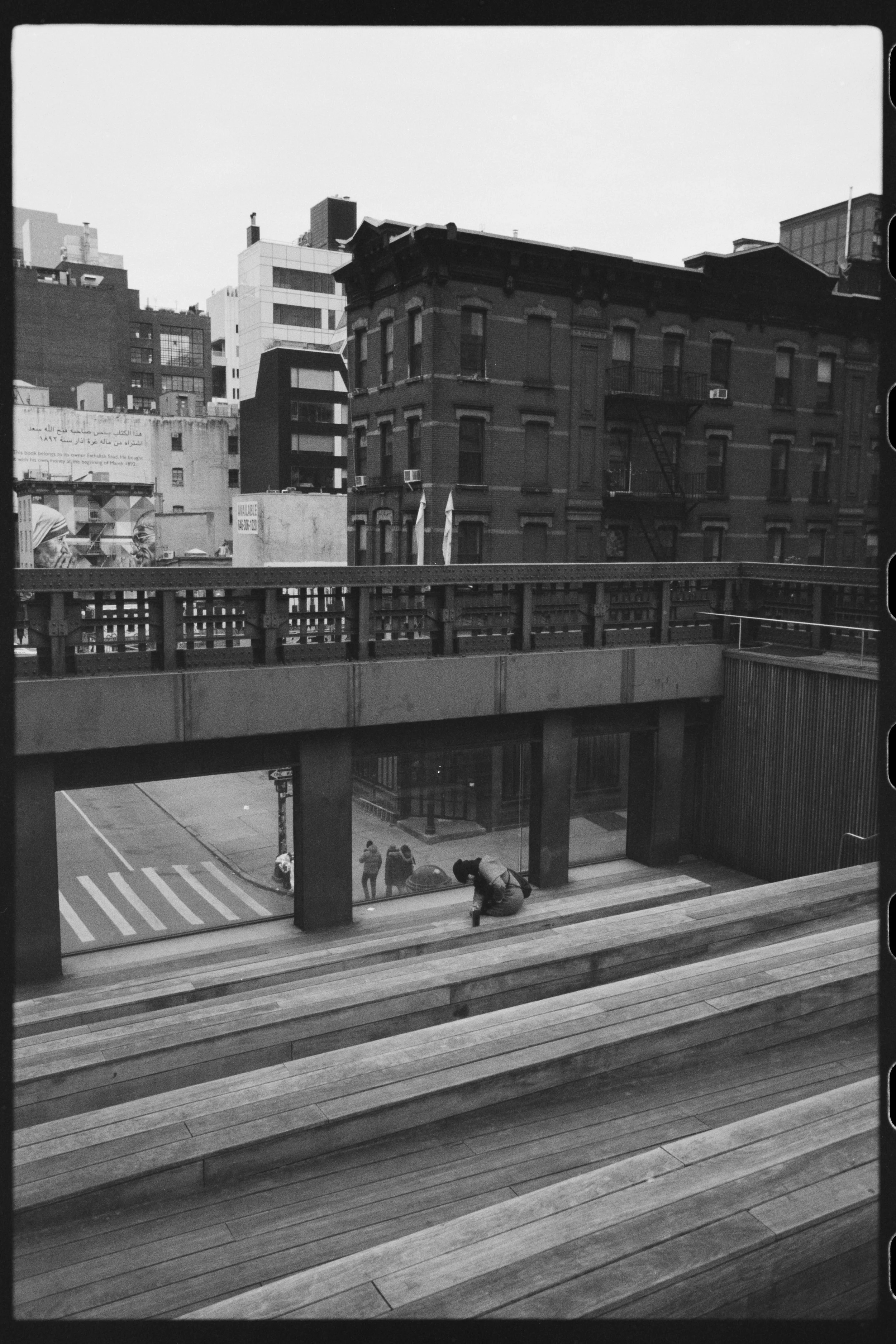 A black and white photo of a city street scene taken from an elevated perspective. In the foreground, a wooden staircase and a sidewalk with three people standing near the door of a building. Two of them are talking while the third is leaning over, p