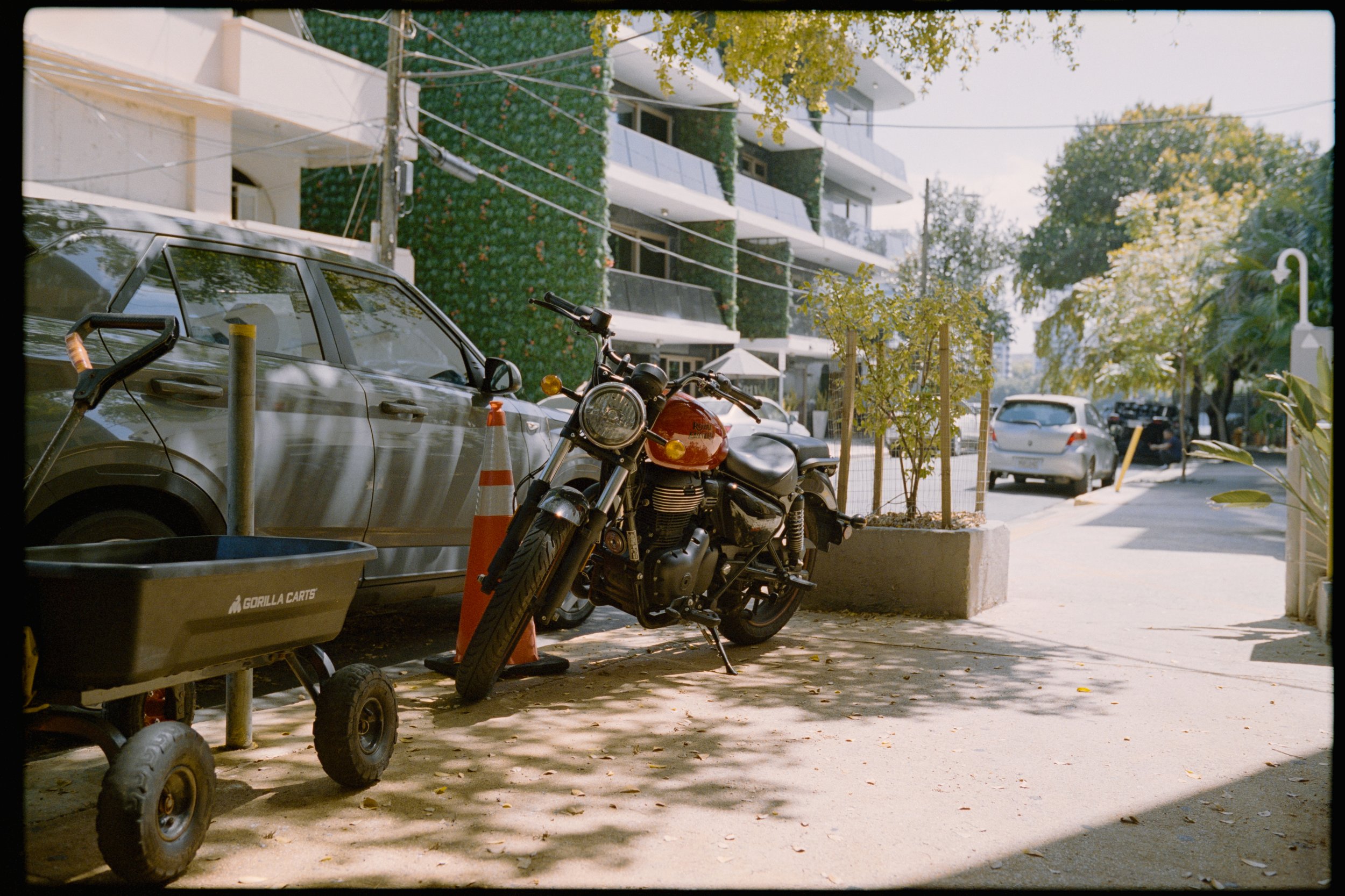 A parking lot with a motorcycle, a gray car, and a container. In the background, there is a multi-story building with balconies and green plant decorations on the facade. Trees with yellow-green leaves are visible, and the scene is illuminated by sun