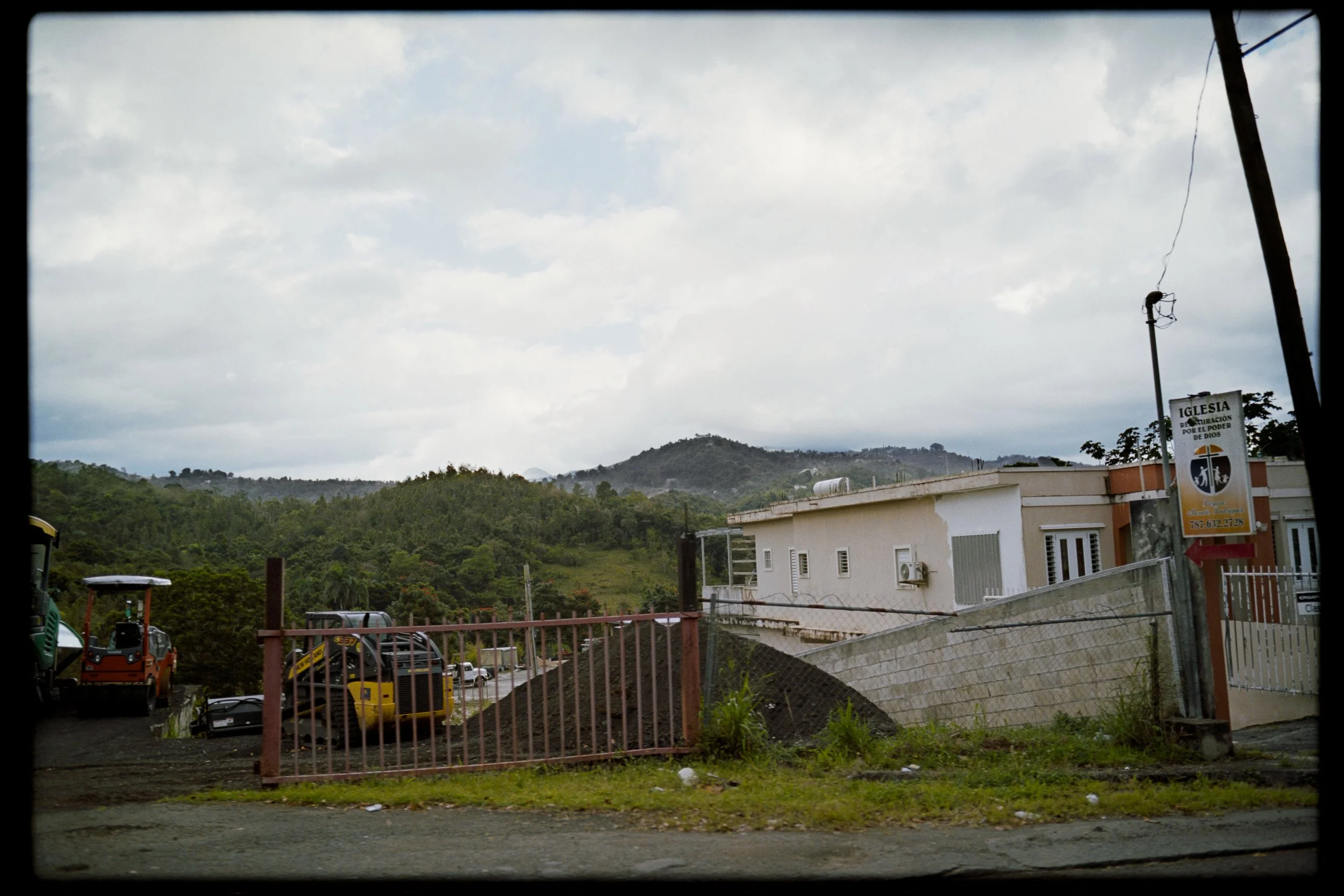 Overcast sky above a residential area with a hillside in the background. Buildings, parked cars, and construction equipment are visible in the foreground, along with a sign for a church or religious center.