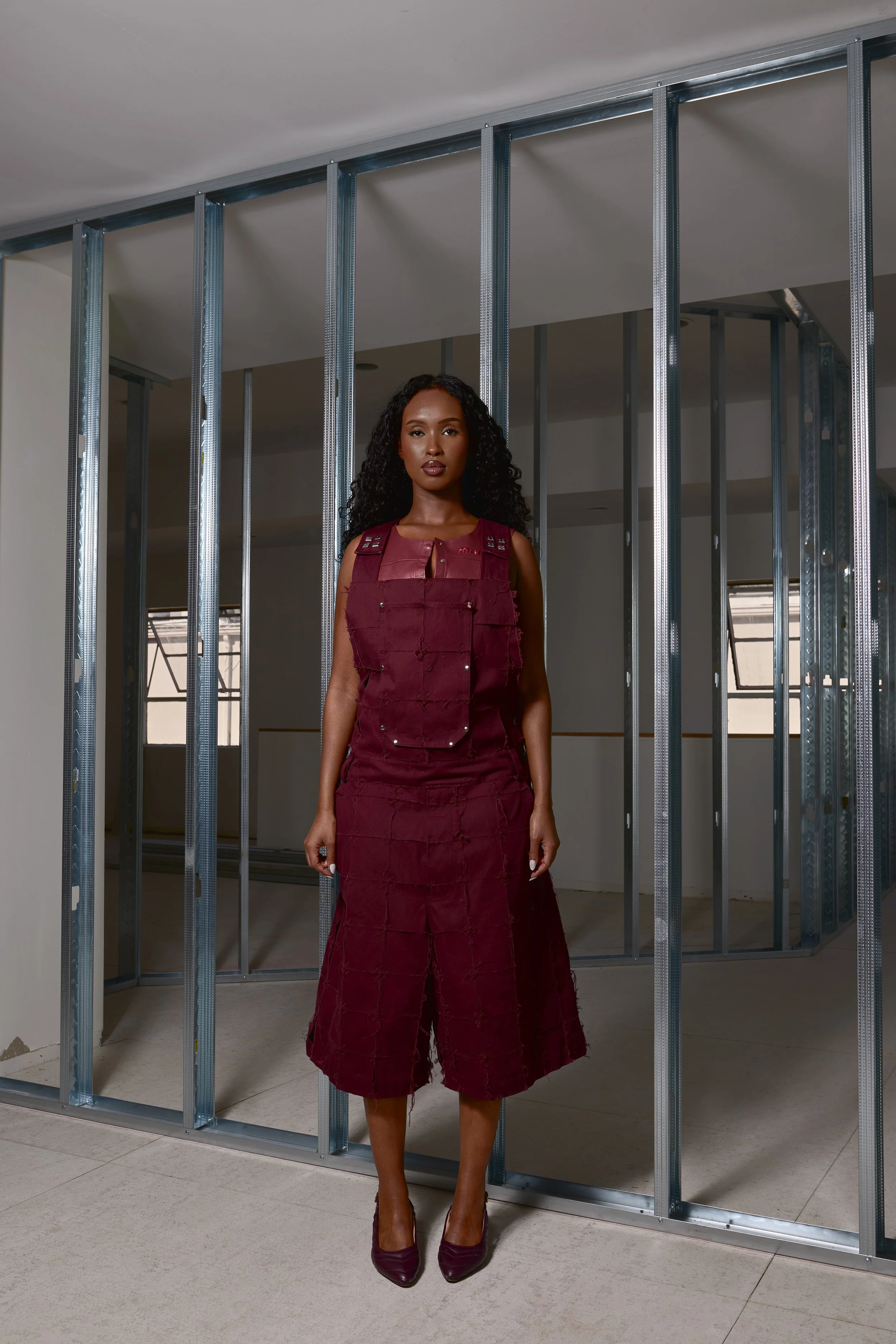 A woman wearing a burgundy outfit featuring a modified vest and wide-legged cropped pants, standing indoors in front of an unfinished wall with metal framing.