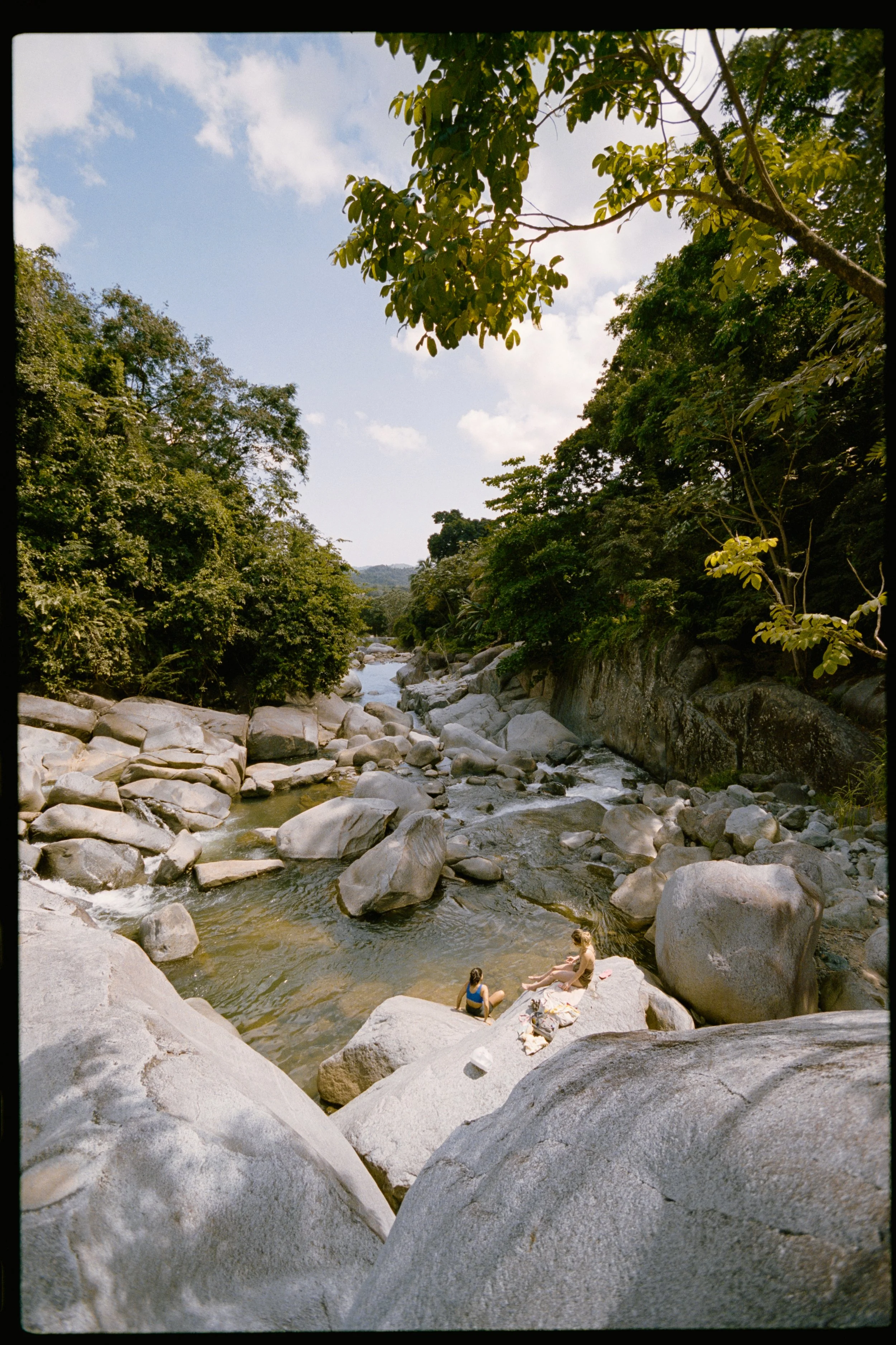 A rocky riverbank with two people relaxing on the rocks near the water, surrounded by lush green trees and a partly cloudy sky.