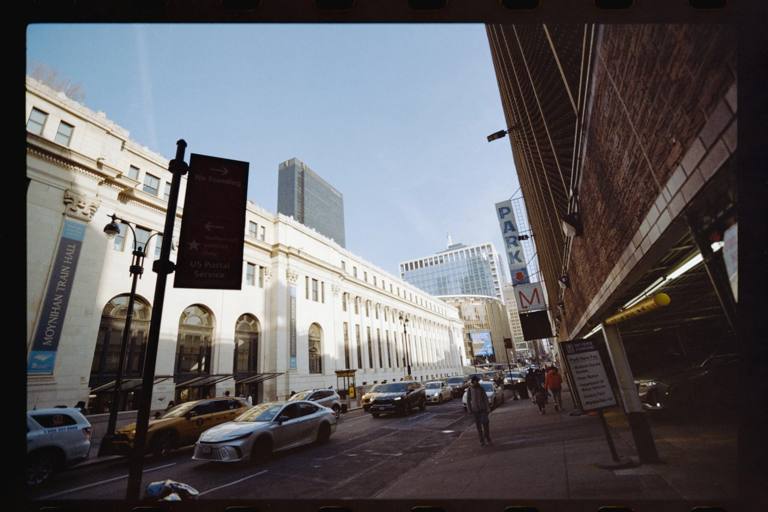City street scene with cars parked and walking pedestrians, tall buildings including a landmark skyscraper, signs indicating parking and restricted areas, and a clear daytime sky.