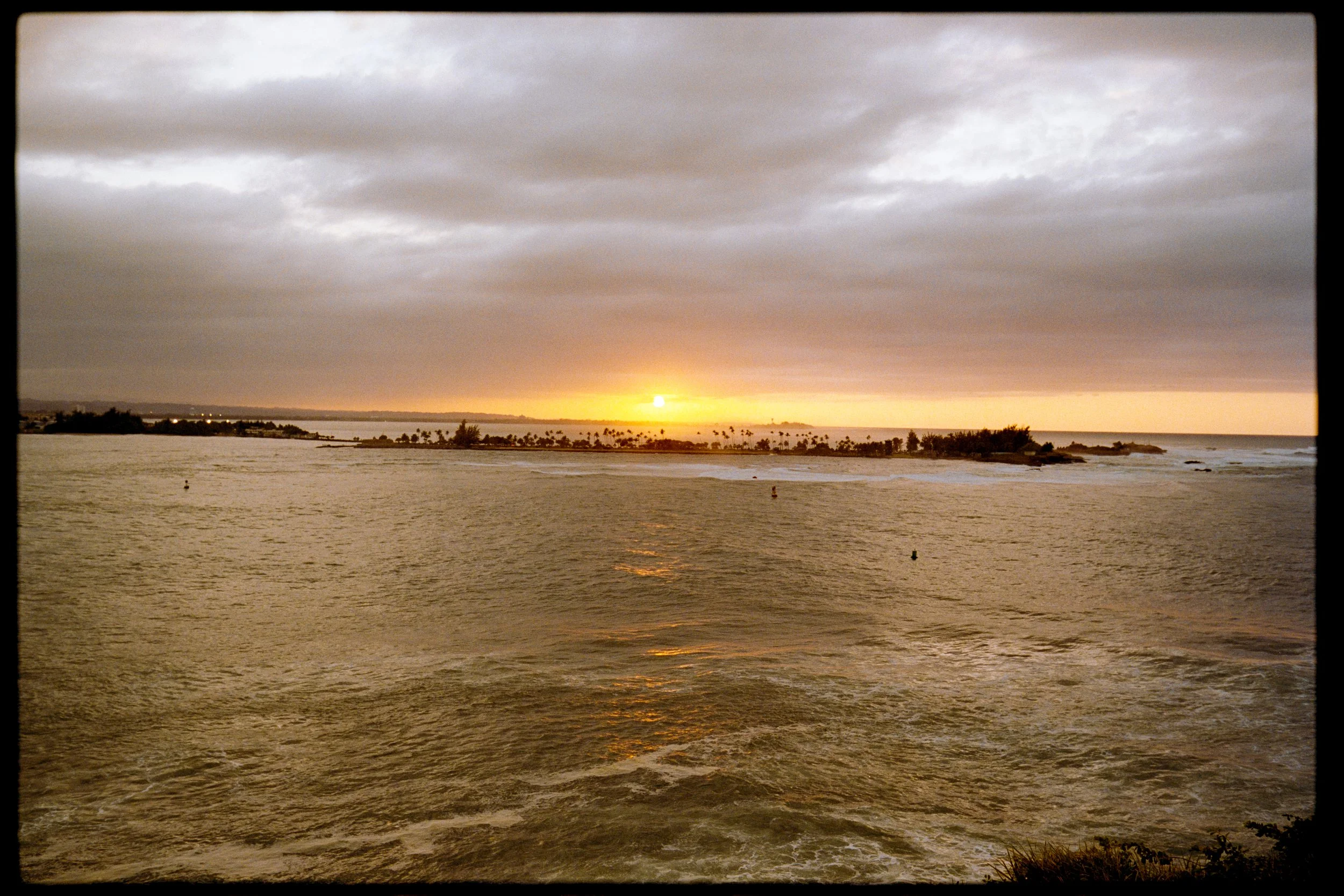 A sunset over a body of water with clouds overhead and a distant shoreline with palm trees.