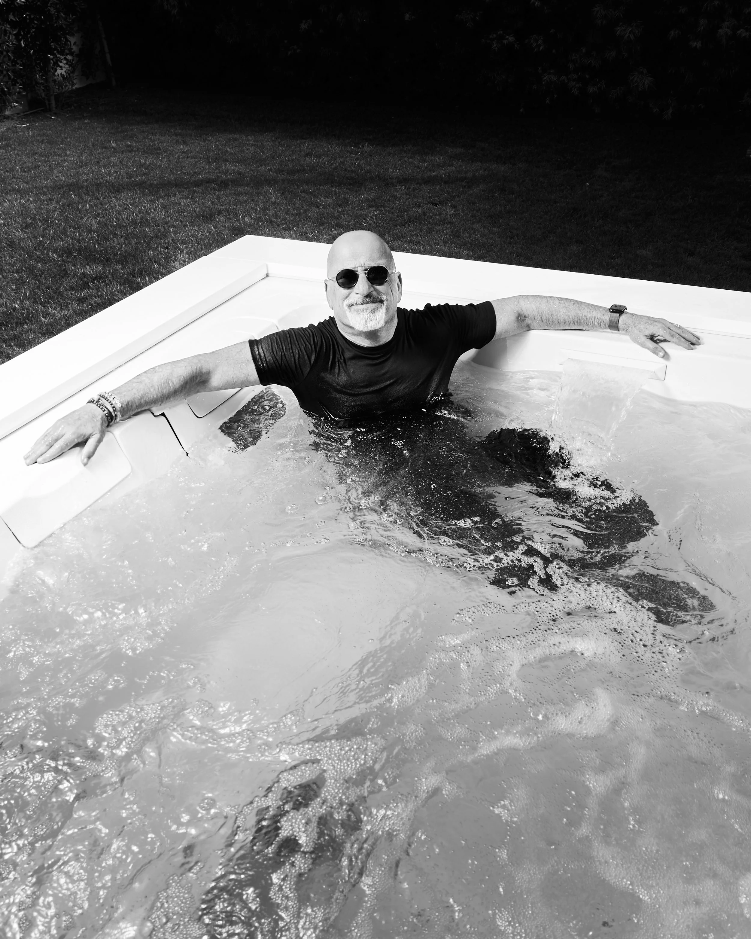A man wearing sunglasses and a black t-shirt is relaxing in a hot tub, smiling with arms resting on the edge, in a backyard at night.