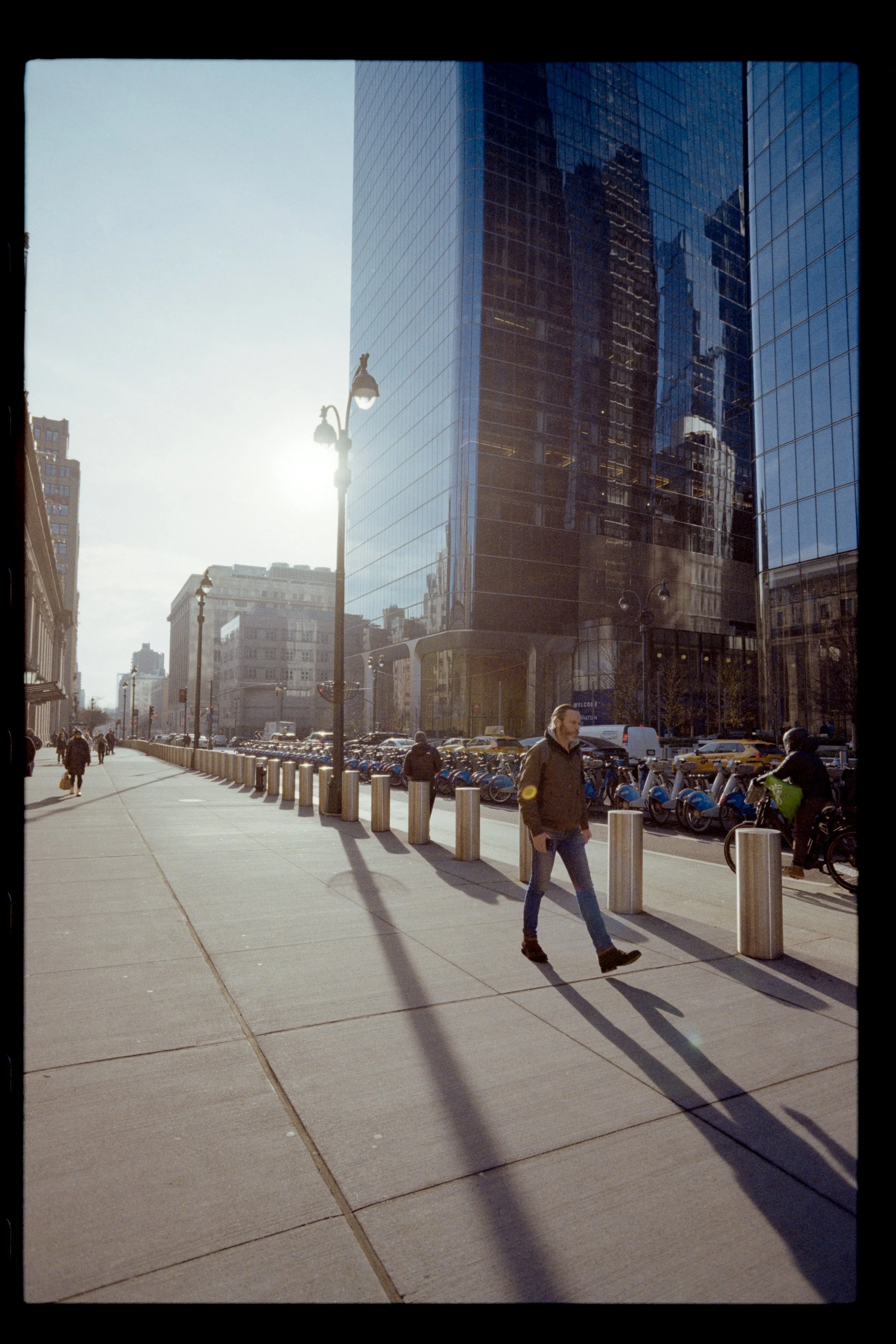City street scene with tall glass skyscraper reflecting surrounding buildings, pedestrians walking, and bike rentals parked along the sidewalk.
