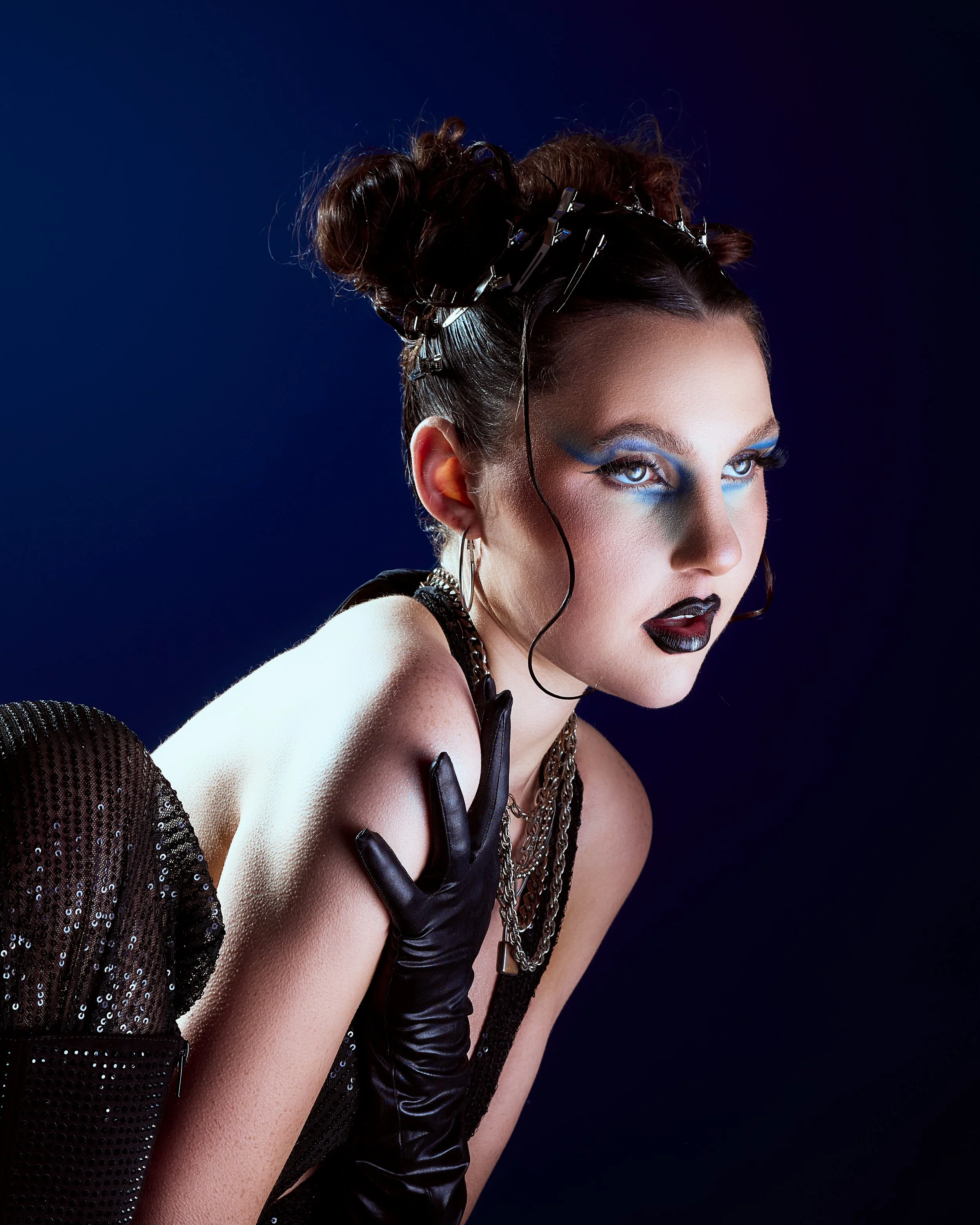 A woman with styled hair, wearing bold makeup with blue eyeshadow and black lipstick, dressed in black with gloves and jewelry, posing against a dark background.