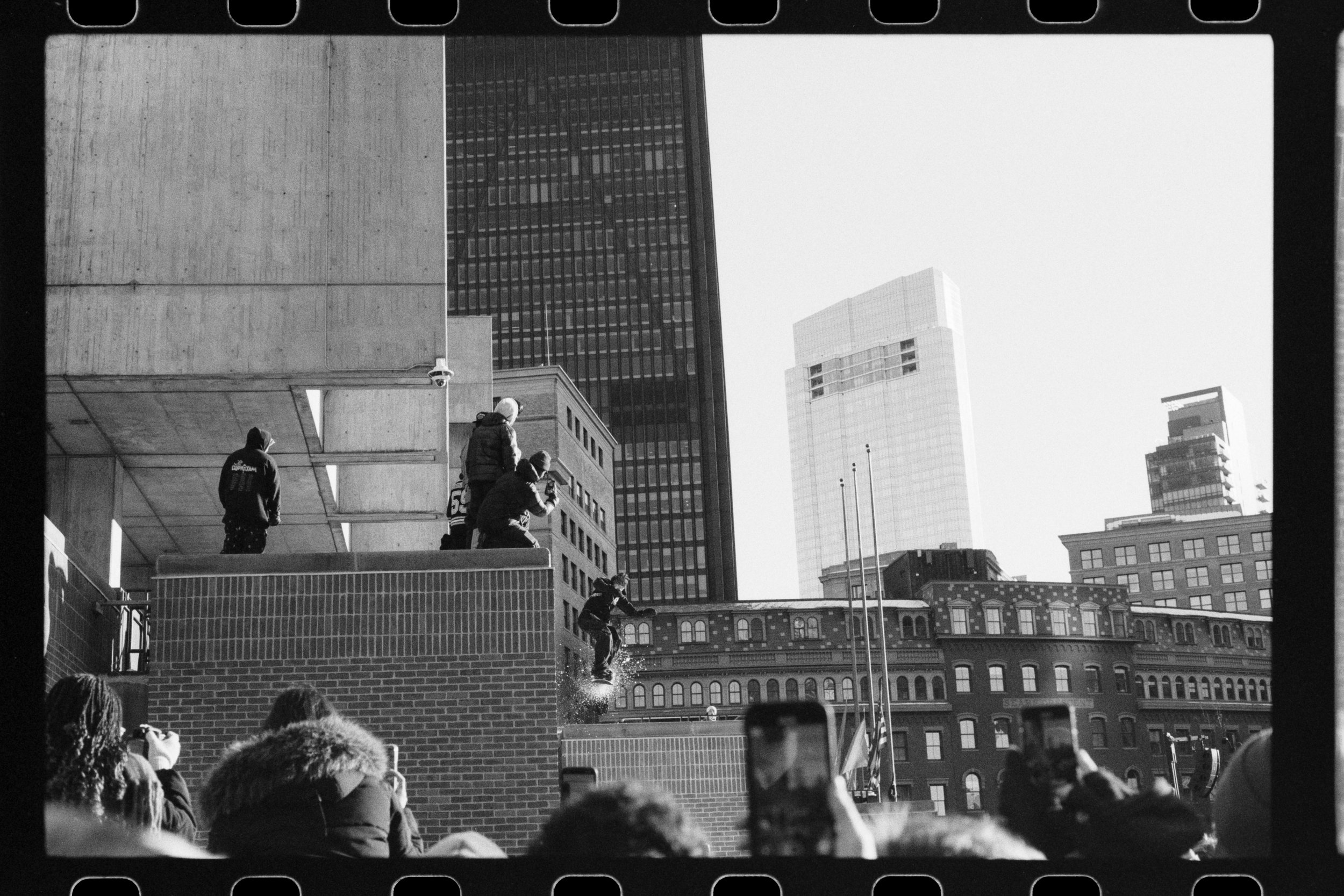 People watching a skateboarder jump off a ledge at an urban skatepark, with tall buildings in the background.