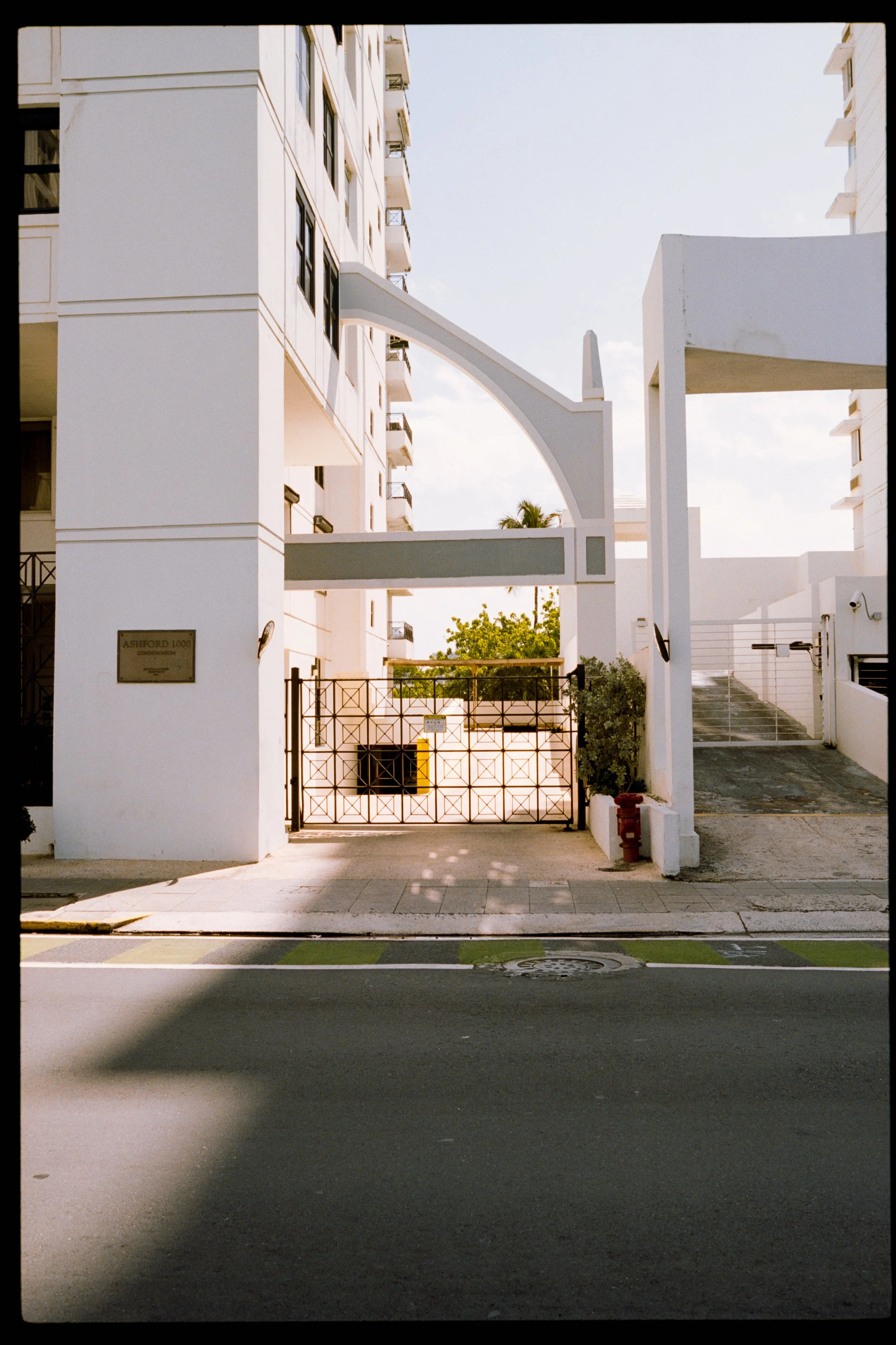 White modern multi-story building with balconies, a gated entrance, and a driveway, set against a partly cloudy sky with a palm tree in the background.