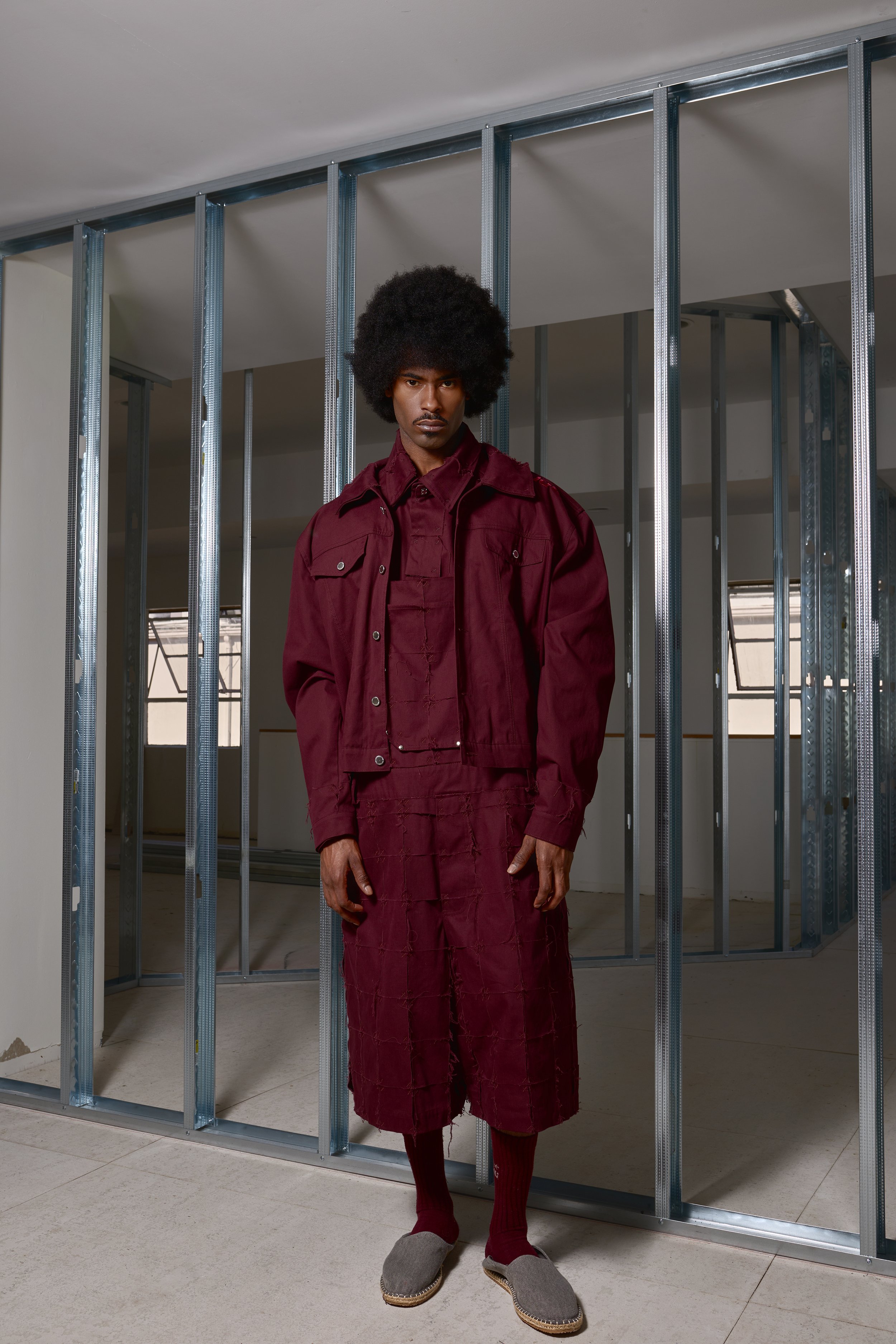 A man with an afro hairstyle wearing a burgundy jacket, shirt, and shorts, standing in front of metal framing in an unfinished room.