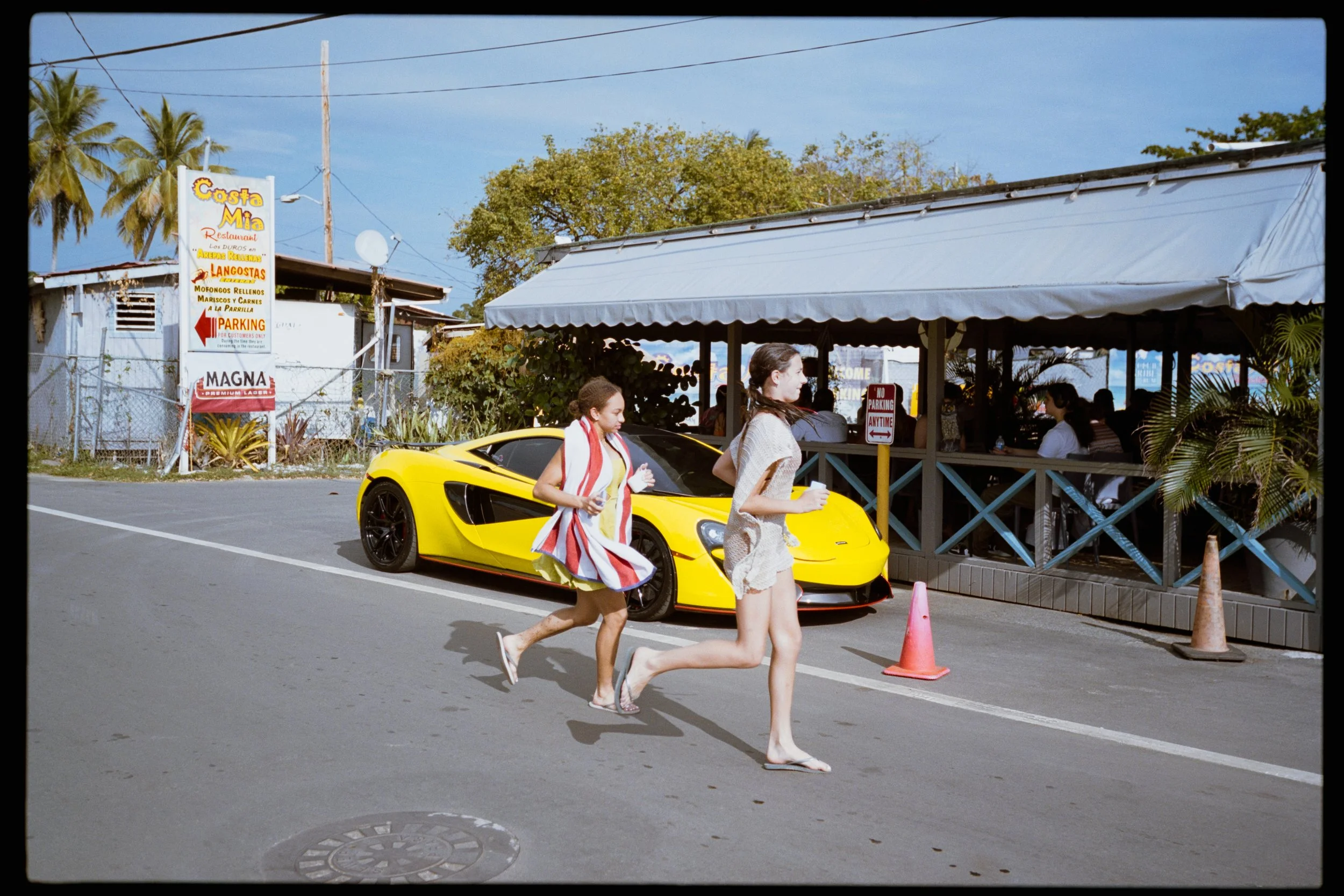 Two women in swimsuits walking on the street beside a yellow sports car outside a restaurant with a covered outdoor seating area. The scene has a tropical vibe with palm trees and a sign for a seafood restaurant.