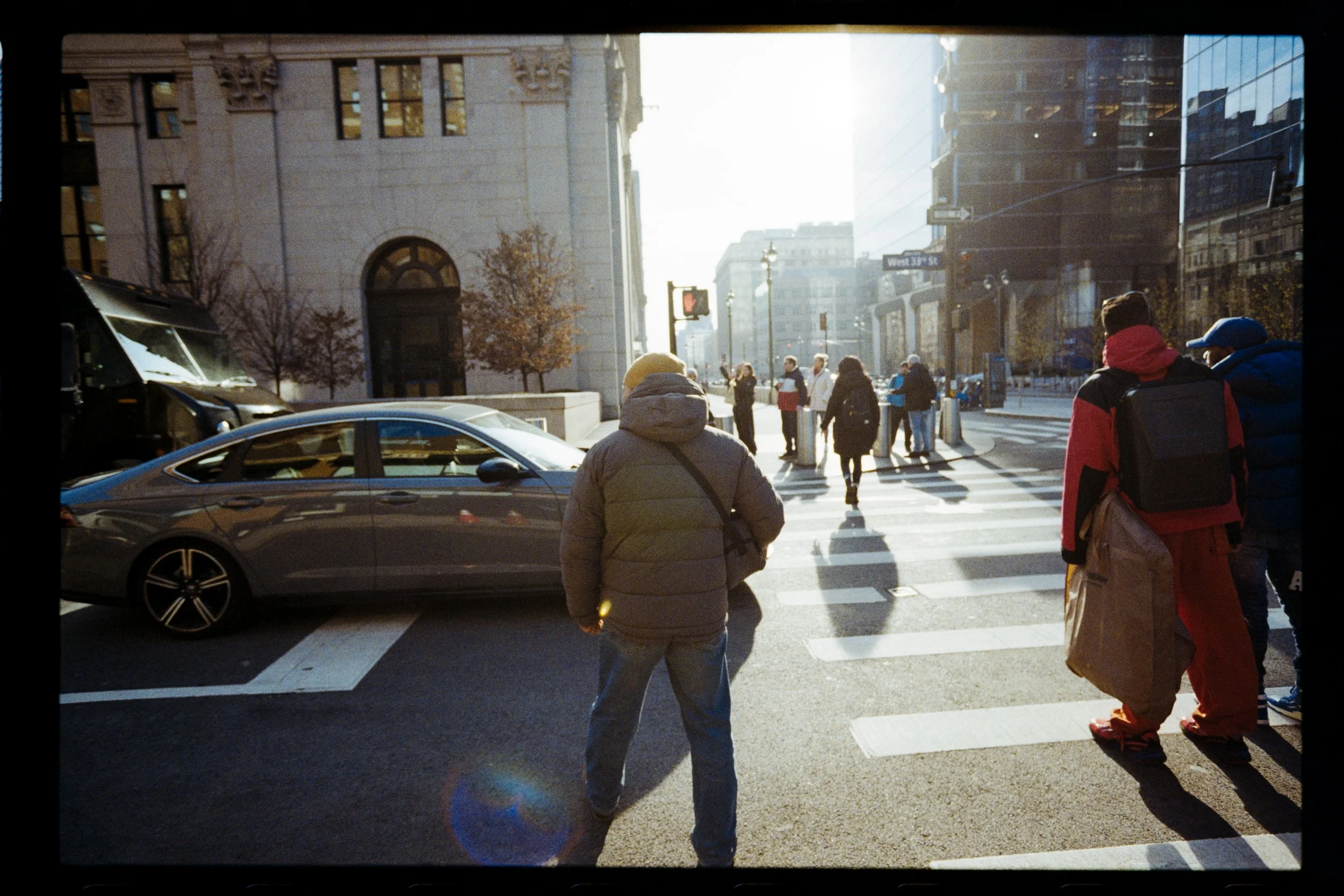 People walking across a city crosswalk during daytime, with a large historic building on the left and modern glass buildings on the right. The sunlight creates backlighting and shadows on the street.