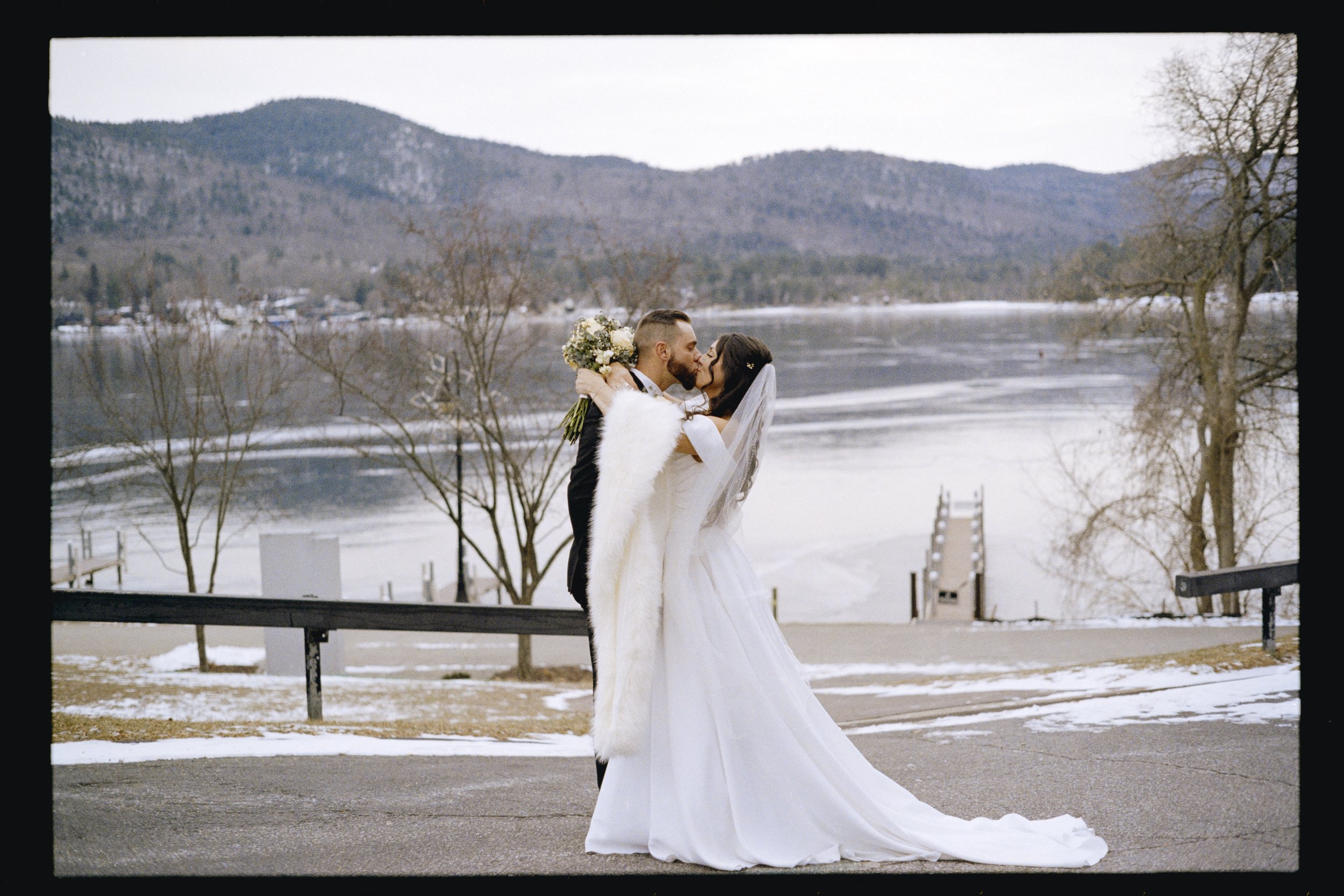 A bride and groom sharing a kiss outdoors by a river, with mountains in the background during winter.