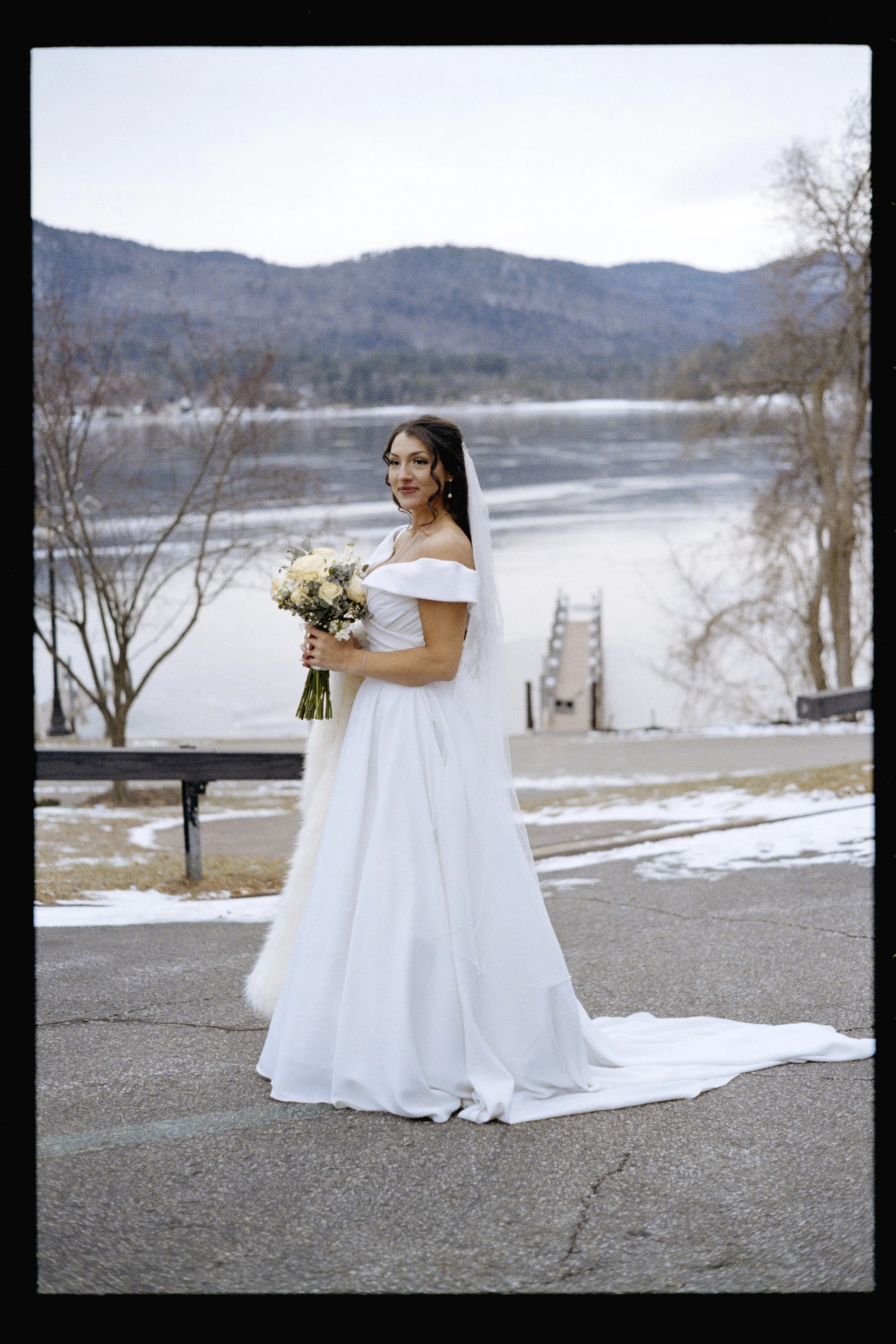 A bride in a white wedding dress holding a bouquet of white roses and greenery stands outdoors near a body of water with mountains in the background on a cold day with some snow on the ground.