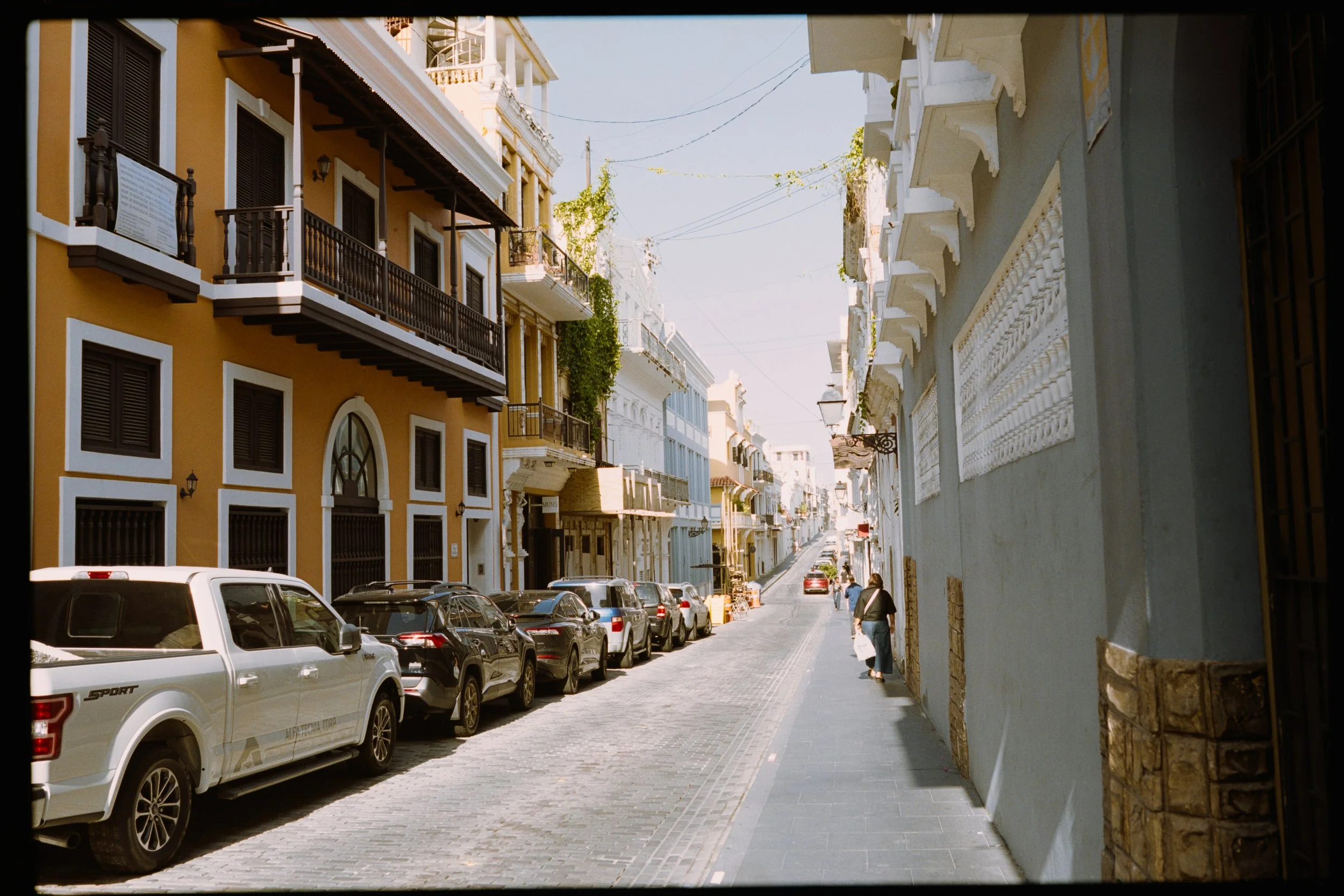 Street view with parked cars on the left and colorful buildings with balconies on the right, under a clear sky.