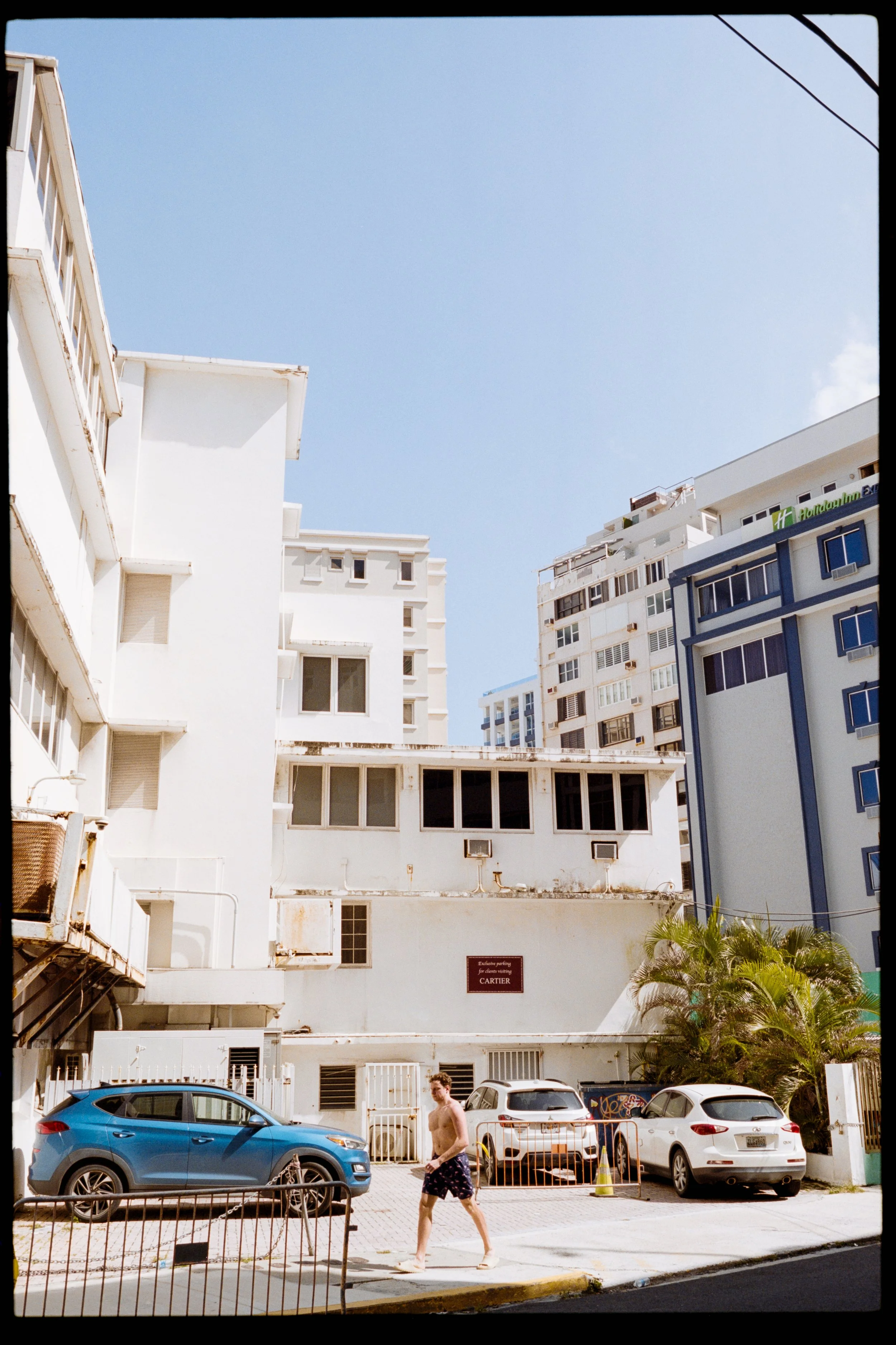 Street view with white and blue buildings, parked cars, palm trees, and a shirtless man walking.