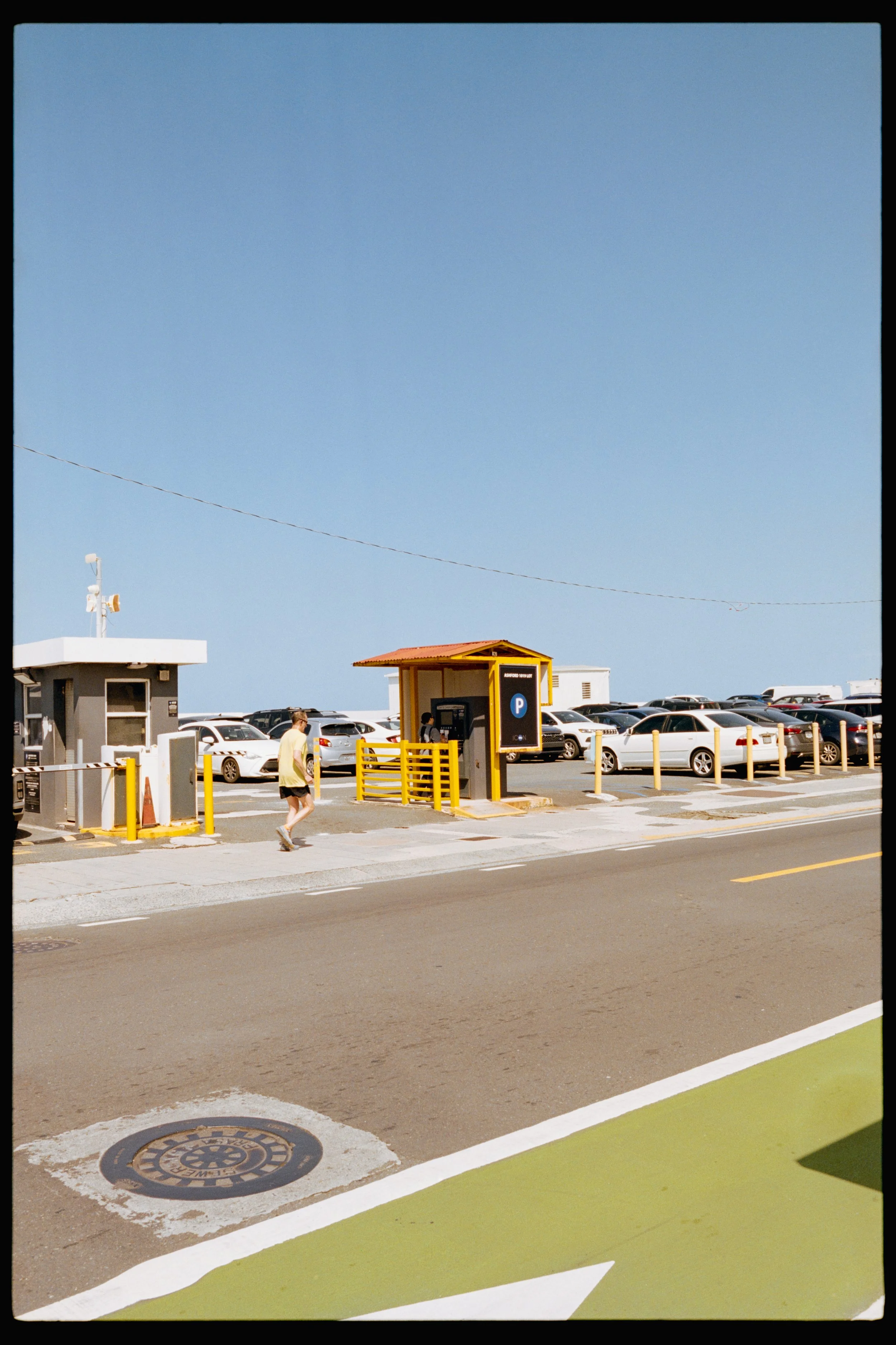 A parking lot with cars parked, a small guard booth, a man walking, and a clear blue sky overhead. There are yellow barriers and a parking payment station.