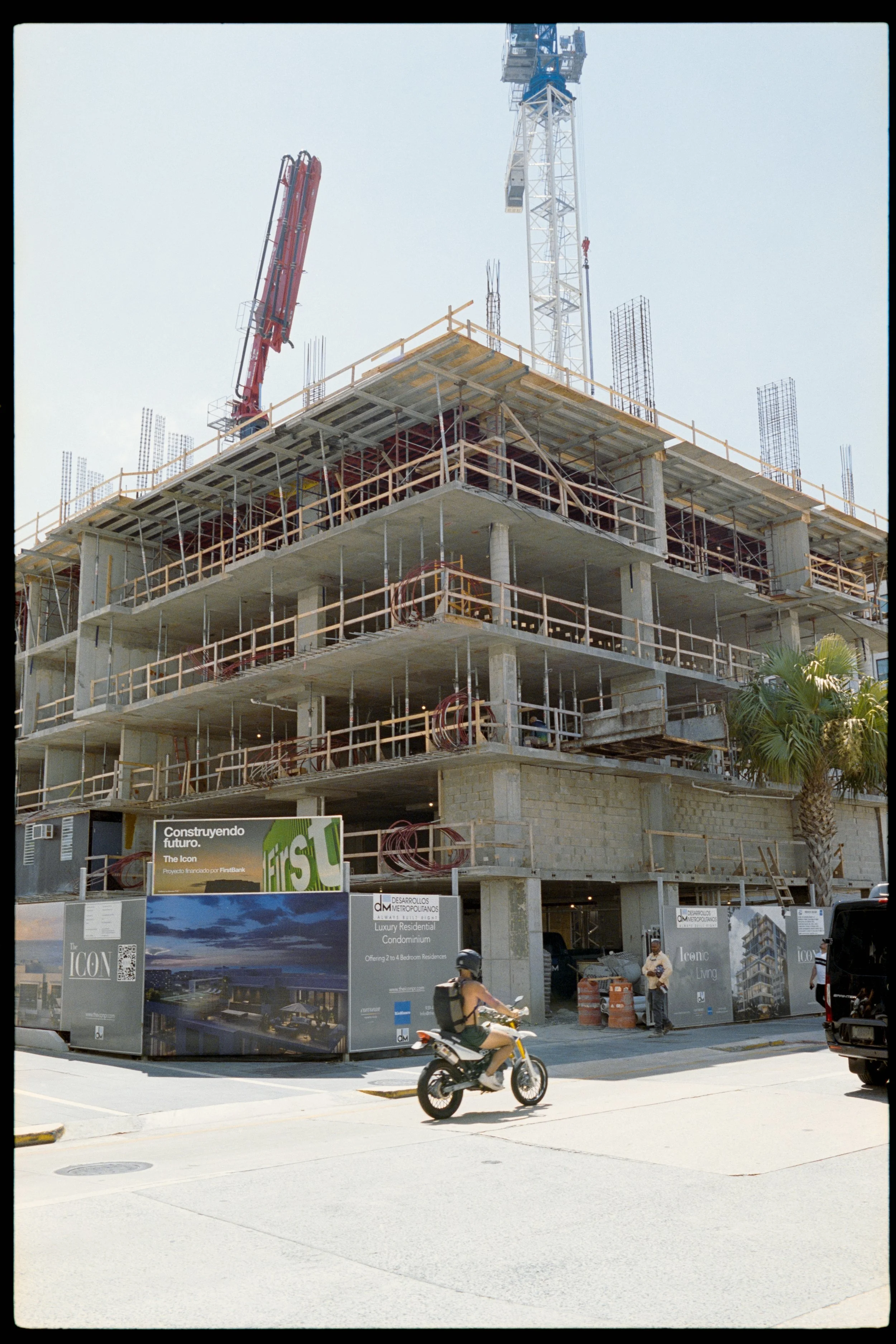 Construction site of a multi-story building with scaffolding and cranes, partially built, with a billboard advertising luxury residential condominiums in front of the street.